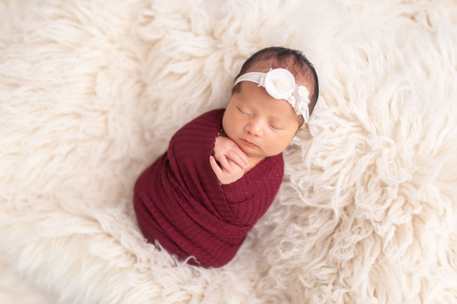 Sleeping newborn wrapped in burgundy fabric on fluffy cream rug.