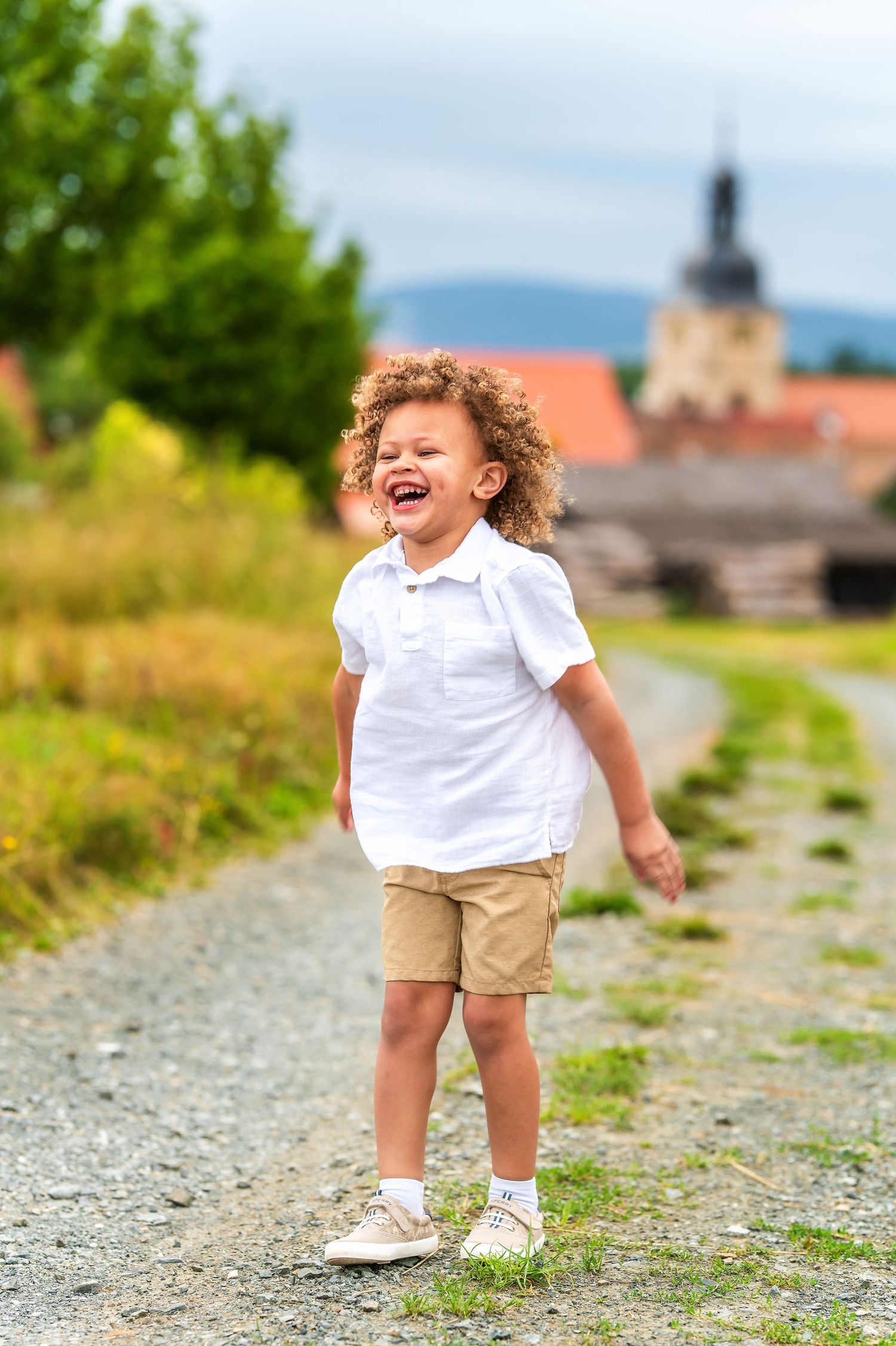 Child playing and laughing on a gravel path with a church spire visible in the rural background.