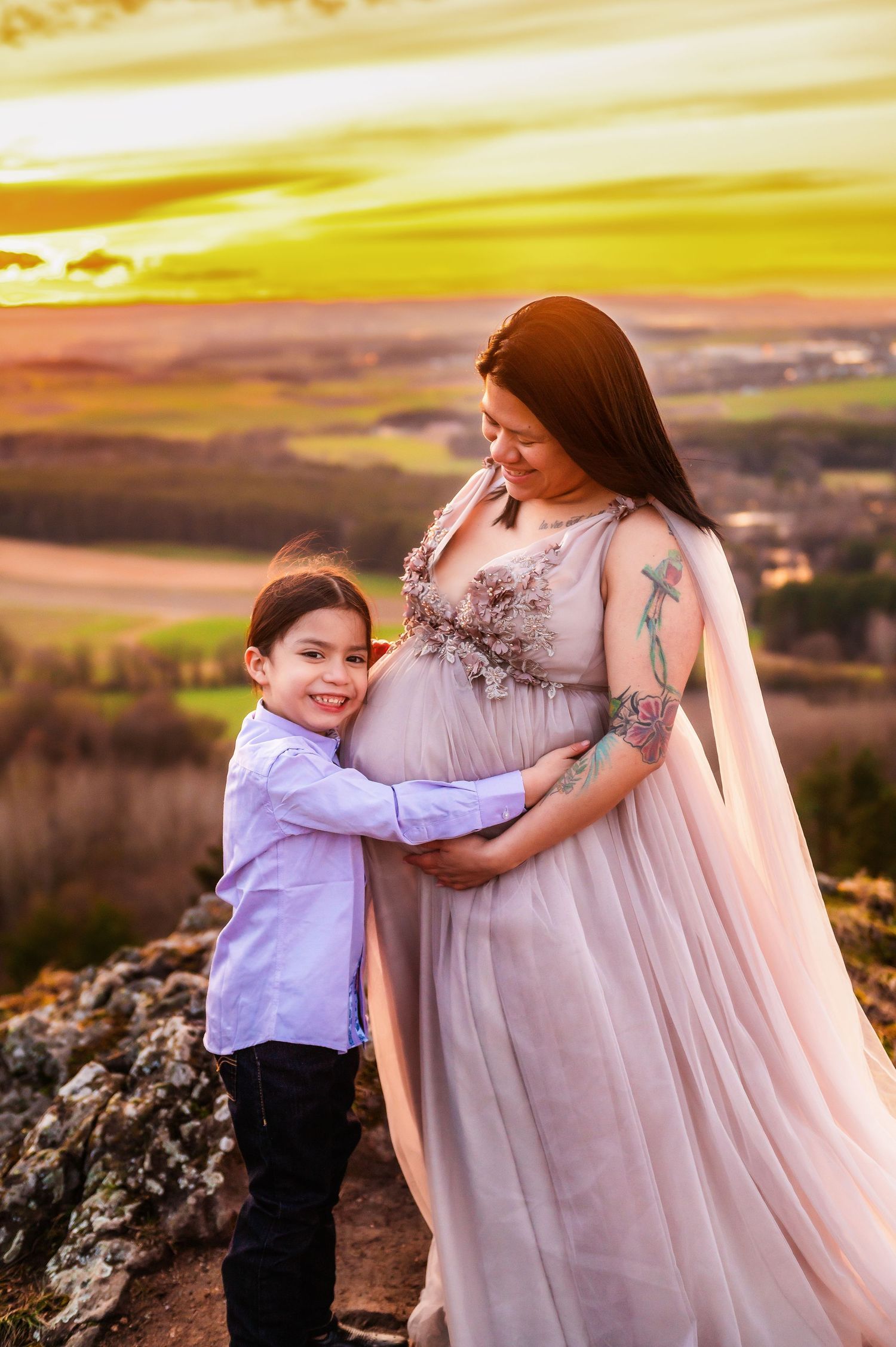 A touching maternity photo at sunset shows a child embracing an expectant mother in a flowing pink dress on a hilltop.