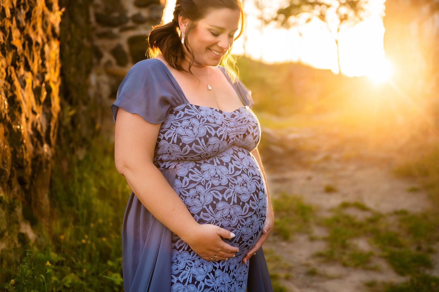 Expectant mother in blue dress poses in golden sunset light while cradling her baby bump in a natural outdoor setting.