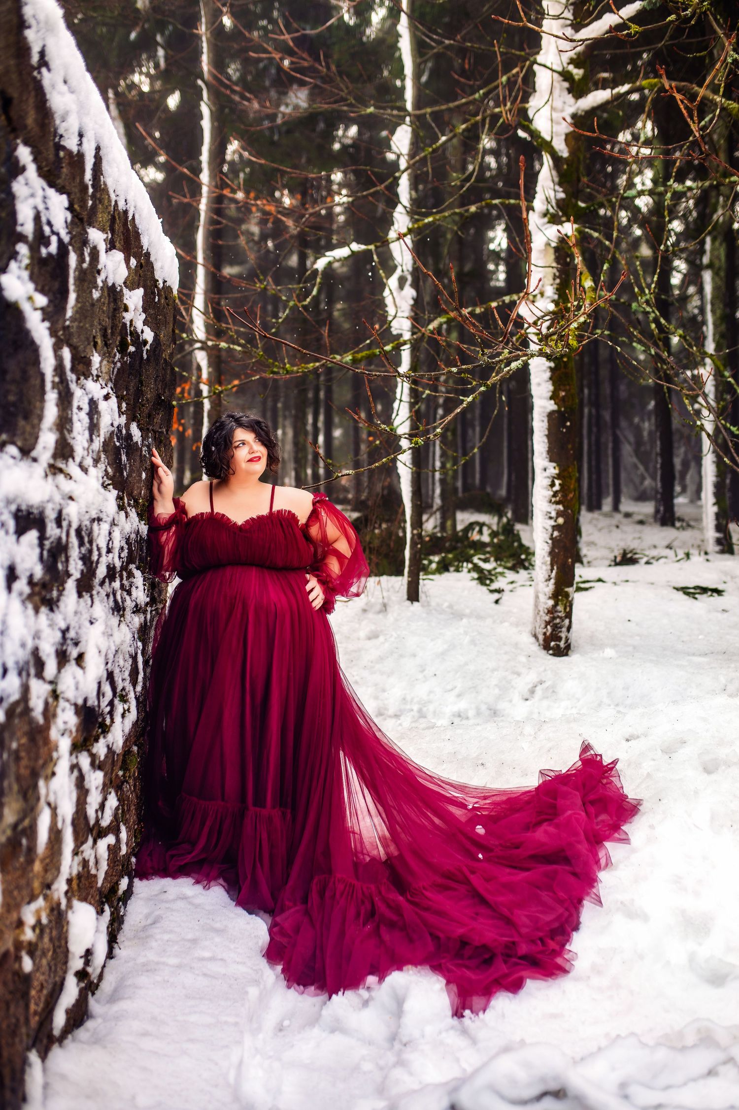 Pregnant woman in red tulle gown poses in snowy forest, leaning against snow-covered tree in winter maternity photo.