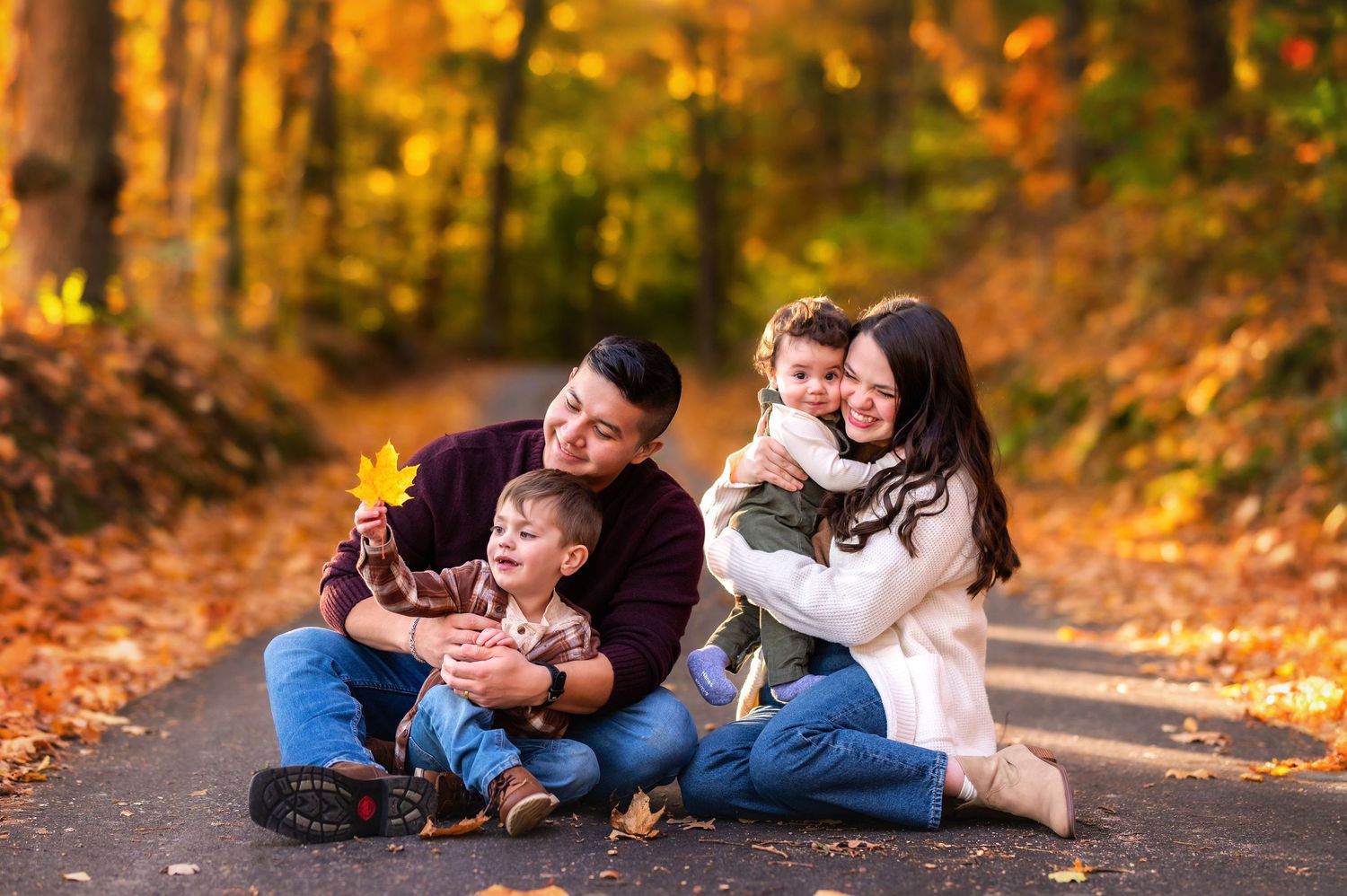 Family of four sitting on a tree-lined path surrounded by vibrant autumn foliage, sharing a joyful moment together.