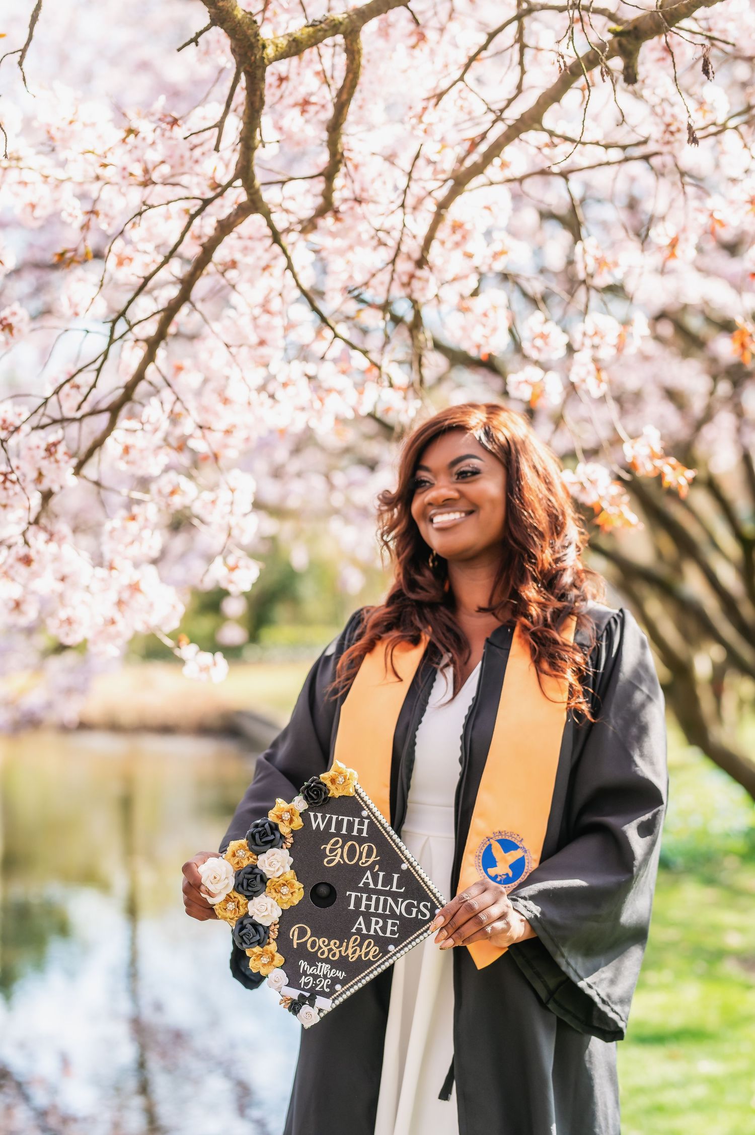 Graduate showing decorated mortarboard cap under flowering cherry trees.