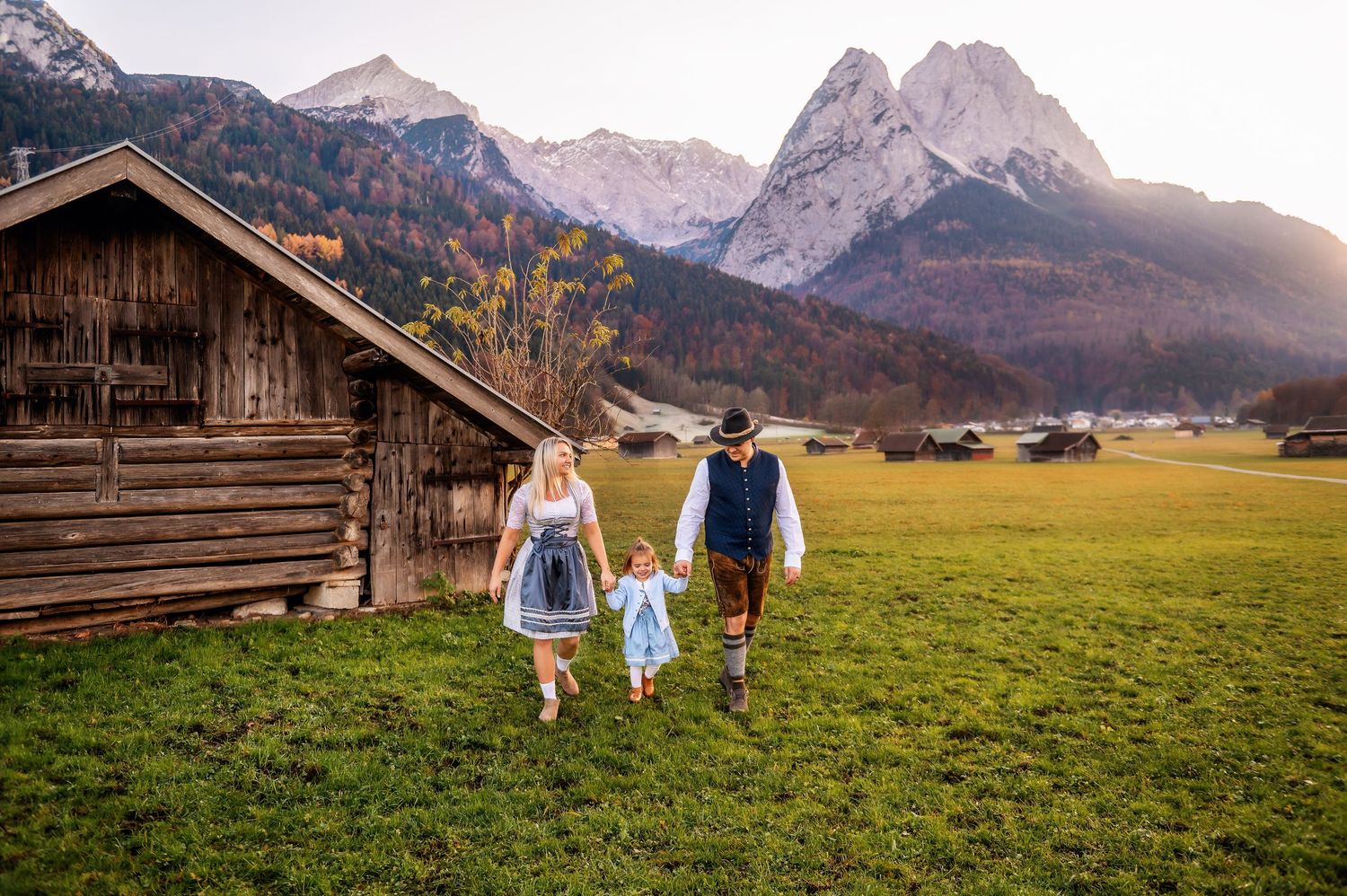 People in traditional Bavarian clothing walk across an alpine meadow with mountains and wooden cabin in the background.