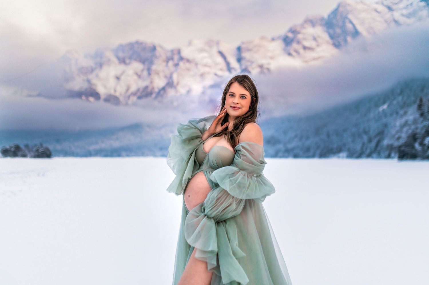 A person in a flowing mint green dress poses on a snowy mountaintop with majestic peaks in the background.