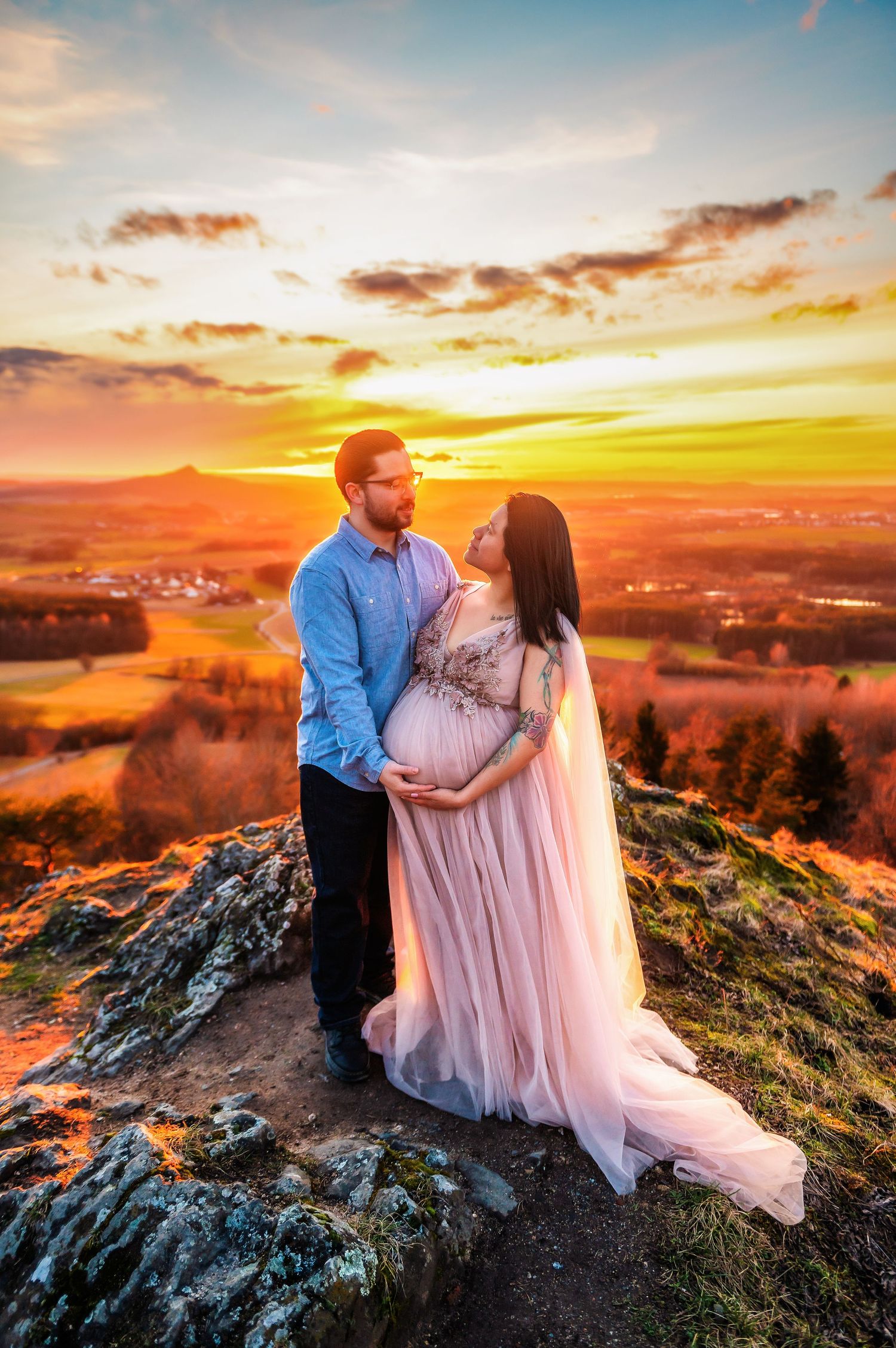 Couple embraces during sunset maternity photoshoot on rocky mountaintop overlook.