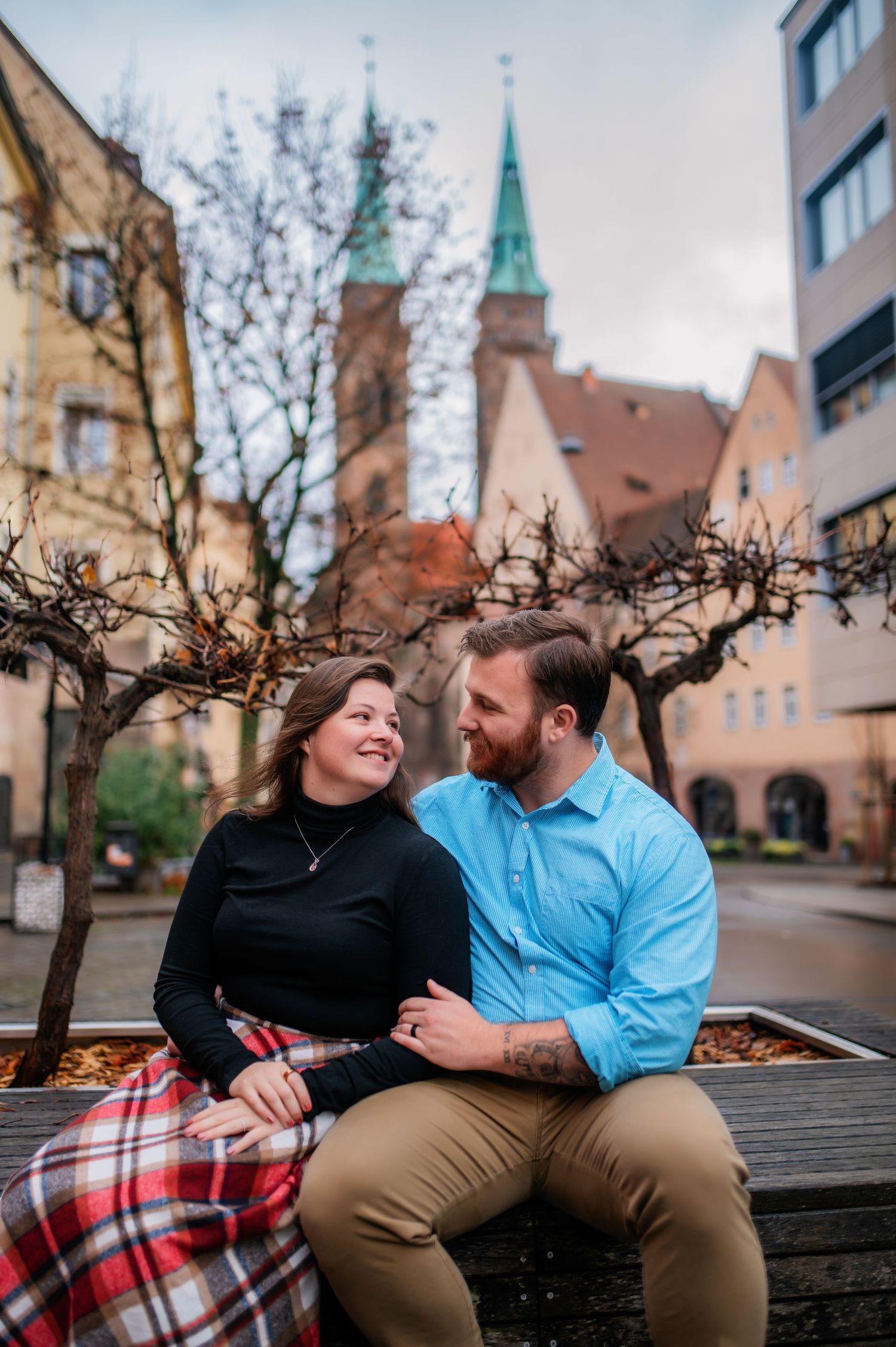 A couple sits together on a bench sharing a cozy plaid blanket in front of historic European architecture.
