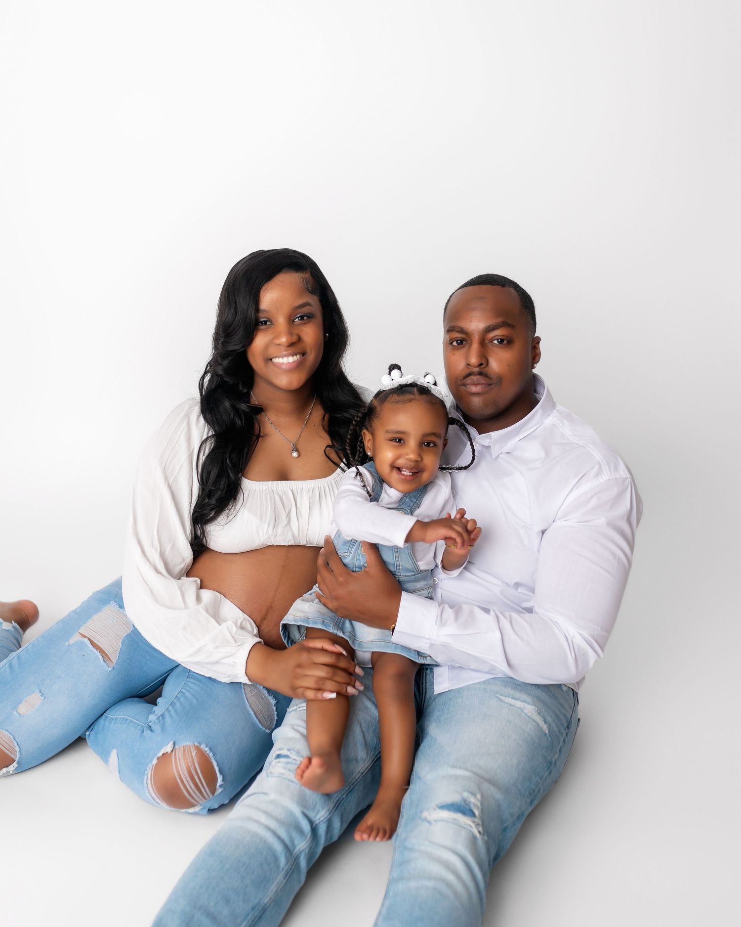 Casual family maternity portrait with denim and white outfits against white backdrop.