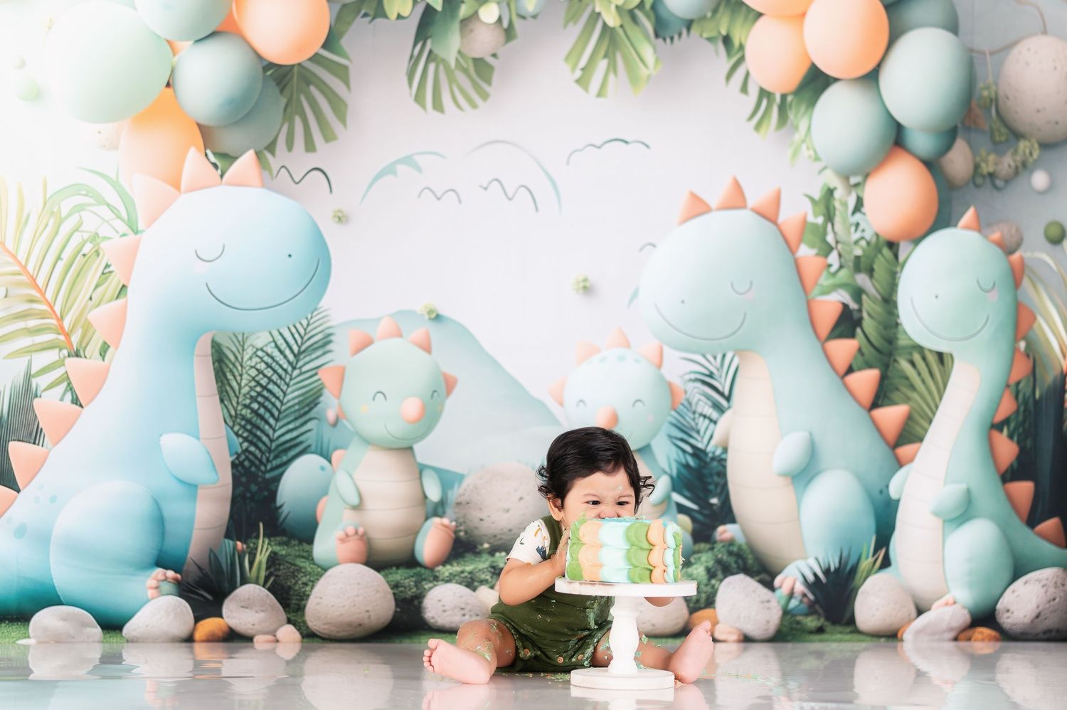 Cute dinosaur themed cake smash setup with pastel blue and peach balloons against white backdrop.