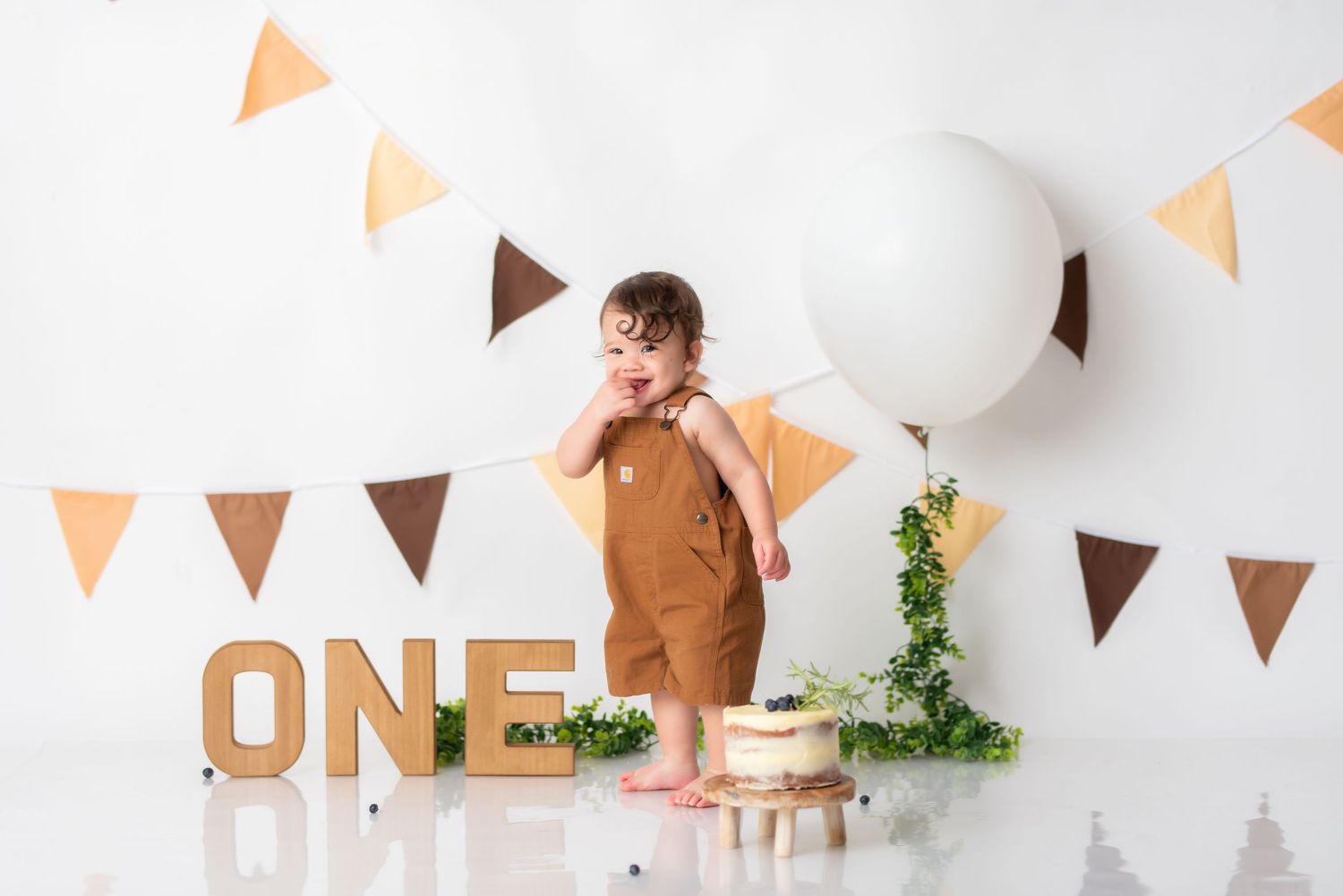 Brown romper cake smash setup with ONE letters and triangular bunting against white background.
