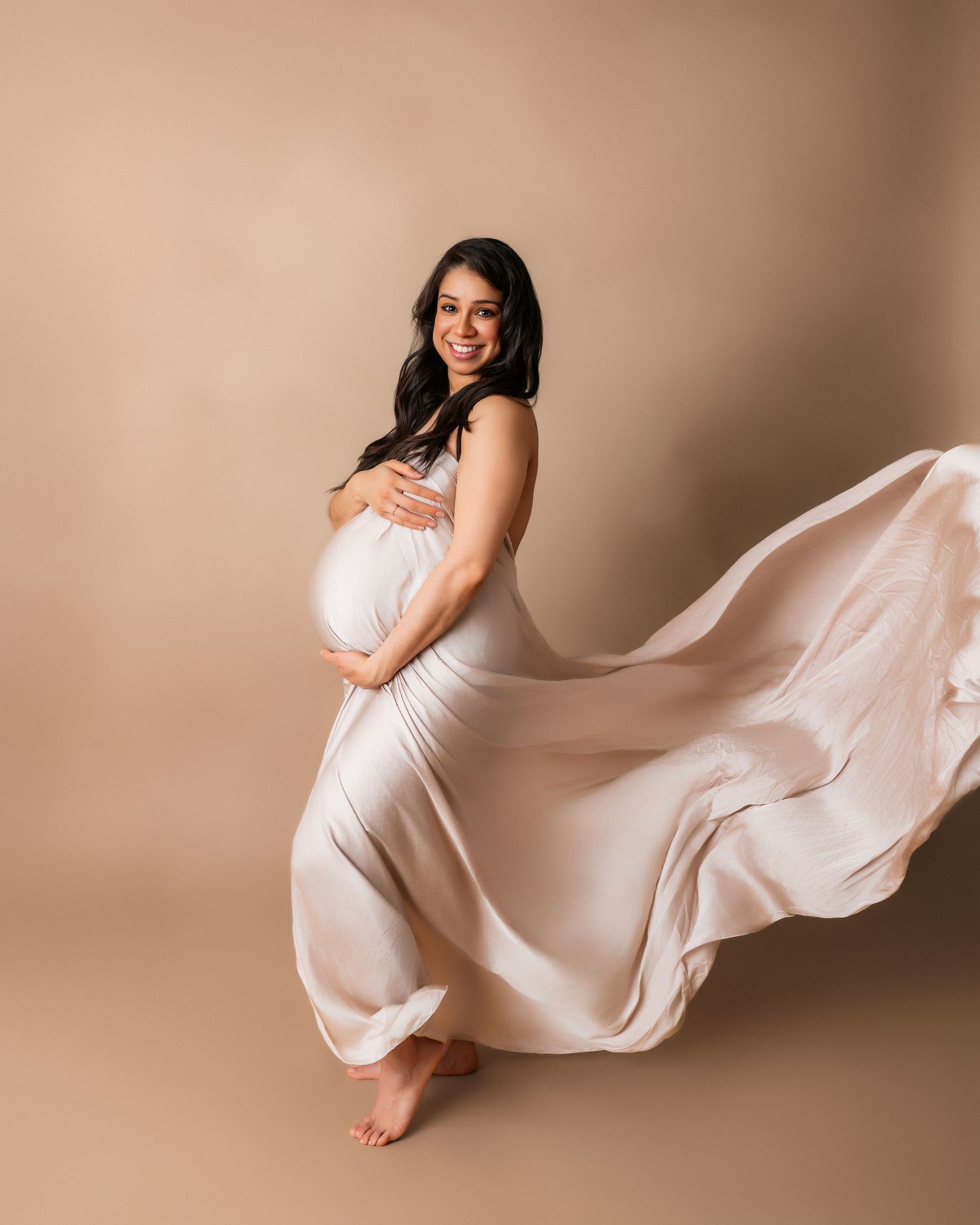 Maternity photo featuring flowing white dress with fabric movement against beige backdrop.