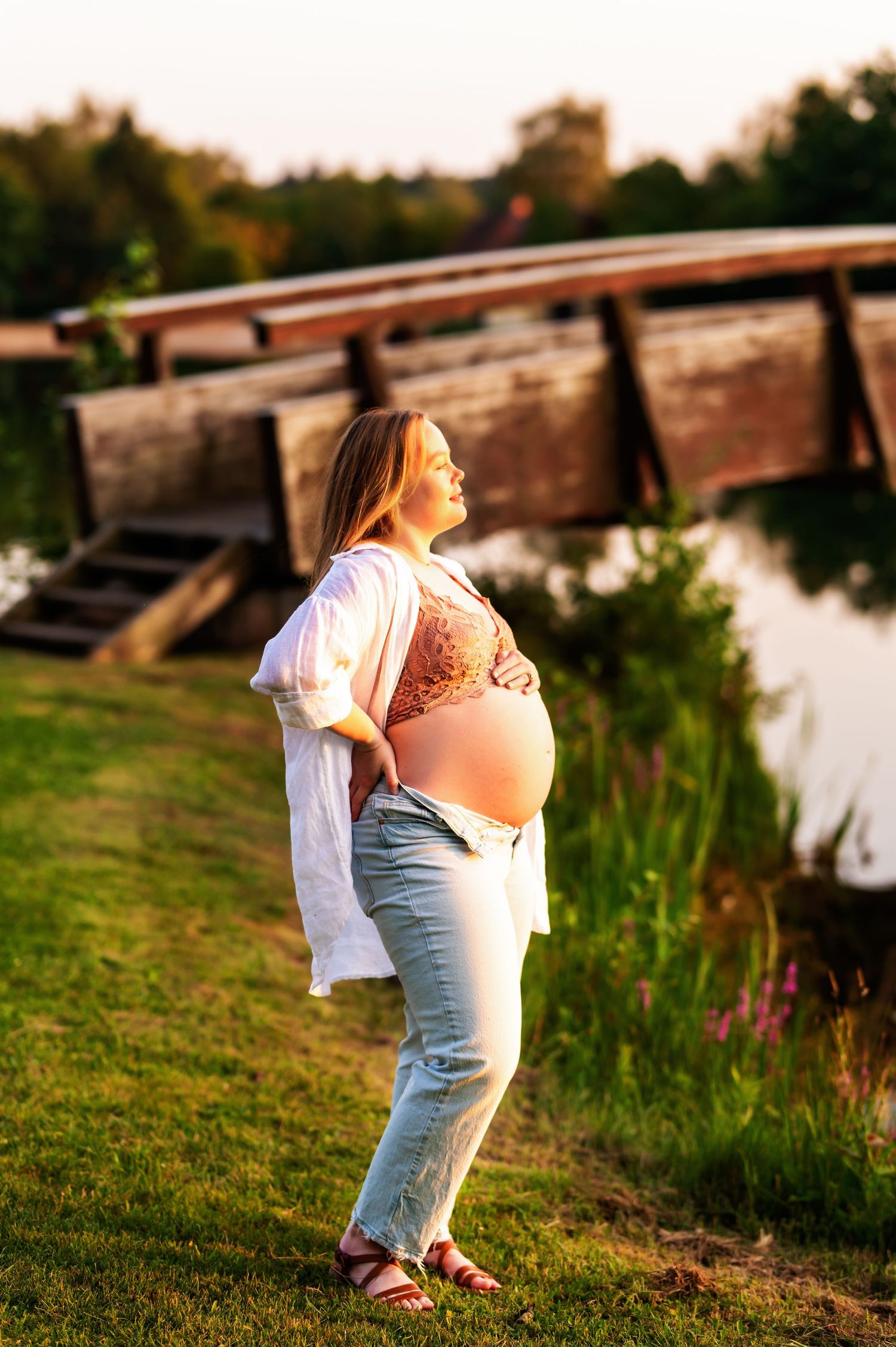 Pregnant woman in white cardigan poses by lake during golden hour maternity session.