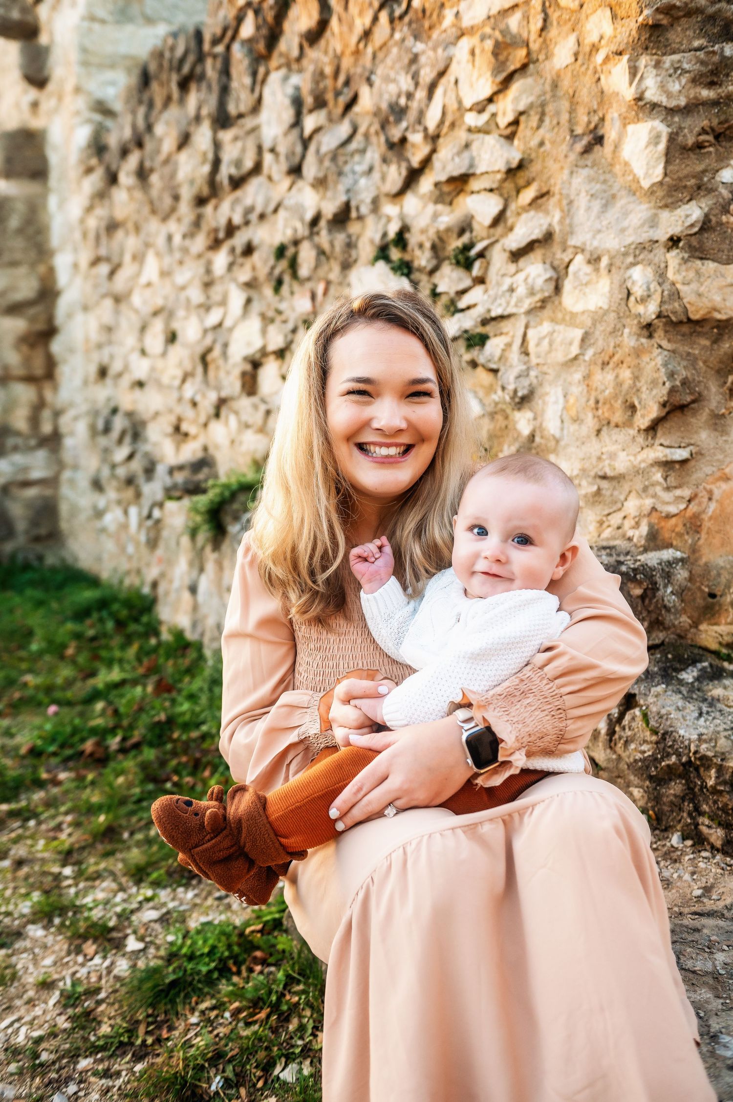 A parent holds a baby while sitting against an old stone wall.
