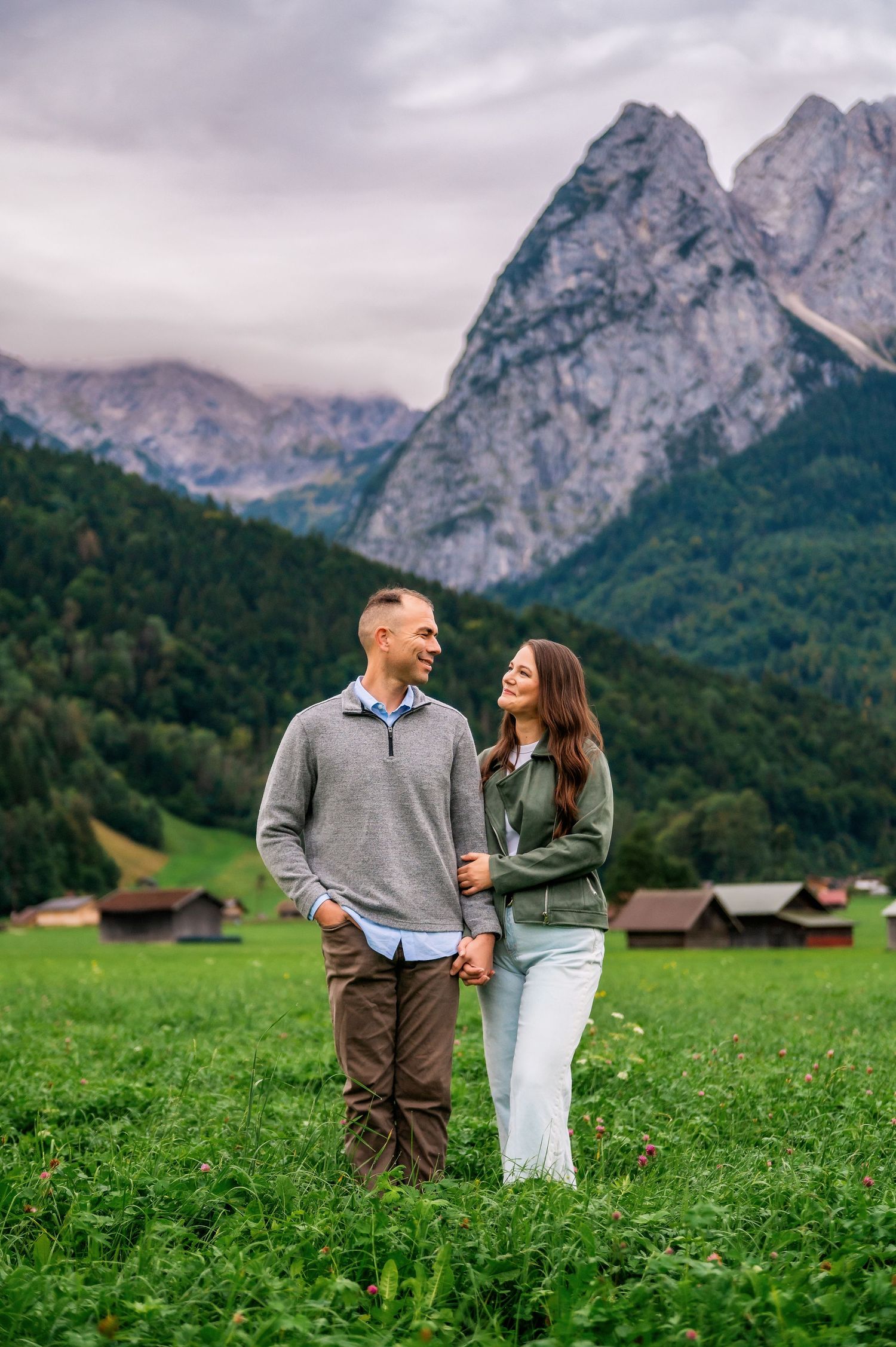 A couple shares a romantic moment in a lush green meadow with dramatic mountain peaks rising in the background.