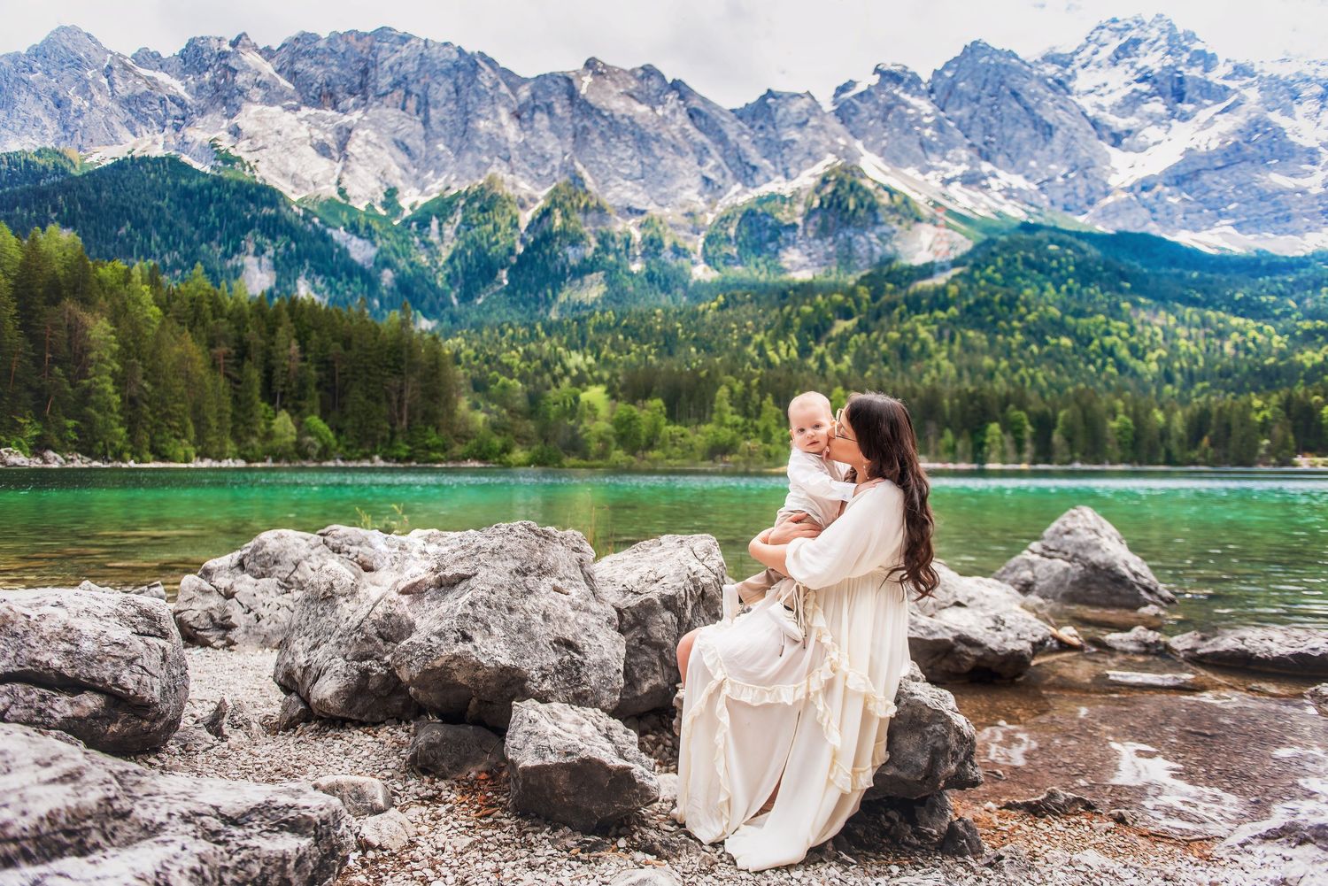 Mother in white dress holds baby on rocky shore of turquoise alpine lake with snow-capped mountains behind.