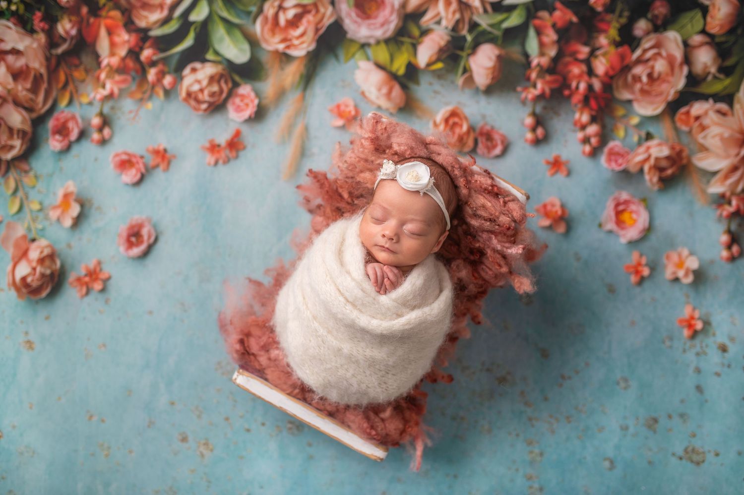 Newborn baby sleeping peacefully wrapped in white, nestled in a small bed surrounded by pink flowers on a blue backdrop.