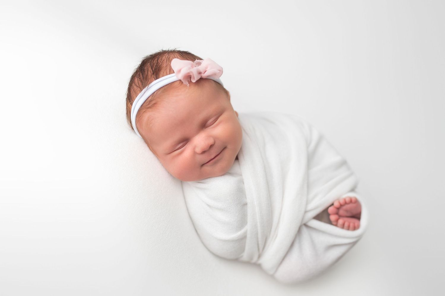 Smiling newborn baby girl wrapped in white swaddle with pink bow headband on white background.