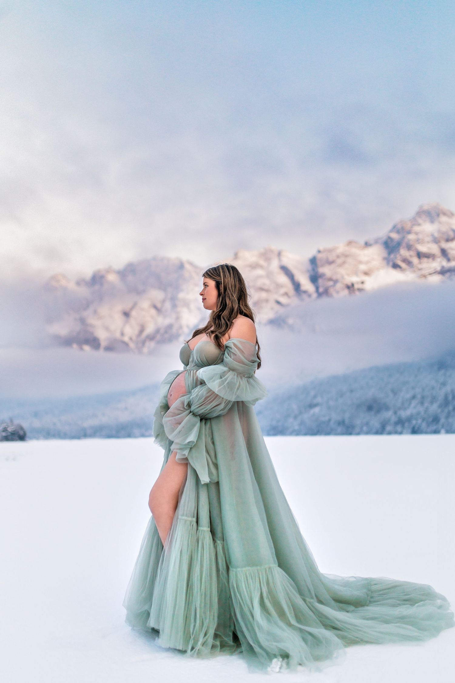 A dreamy maternity shoot in a flowing mint green dress against a snowy mountain landscape at dusk.