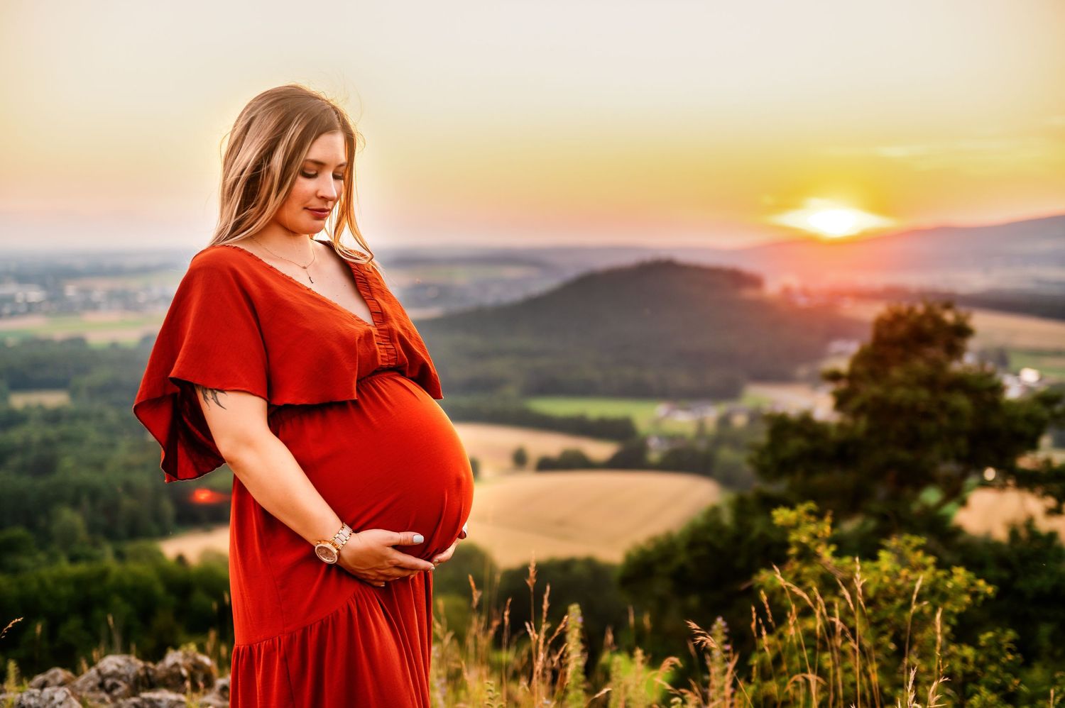 Pregnant woman in red dress stands on hilltop during dramatic sunset maternity photo.