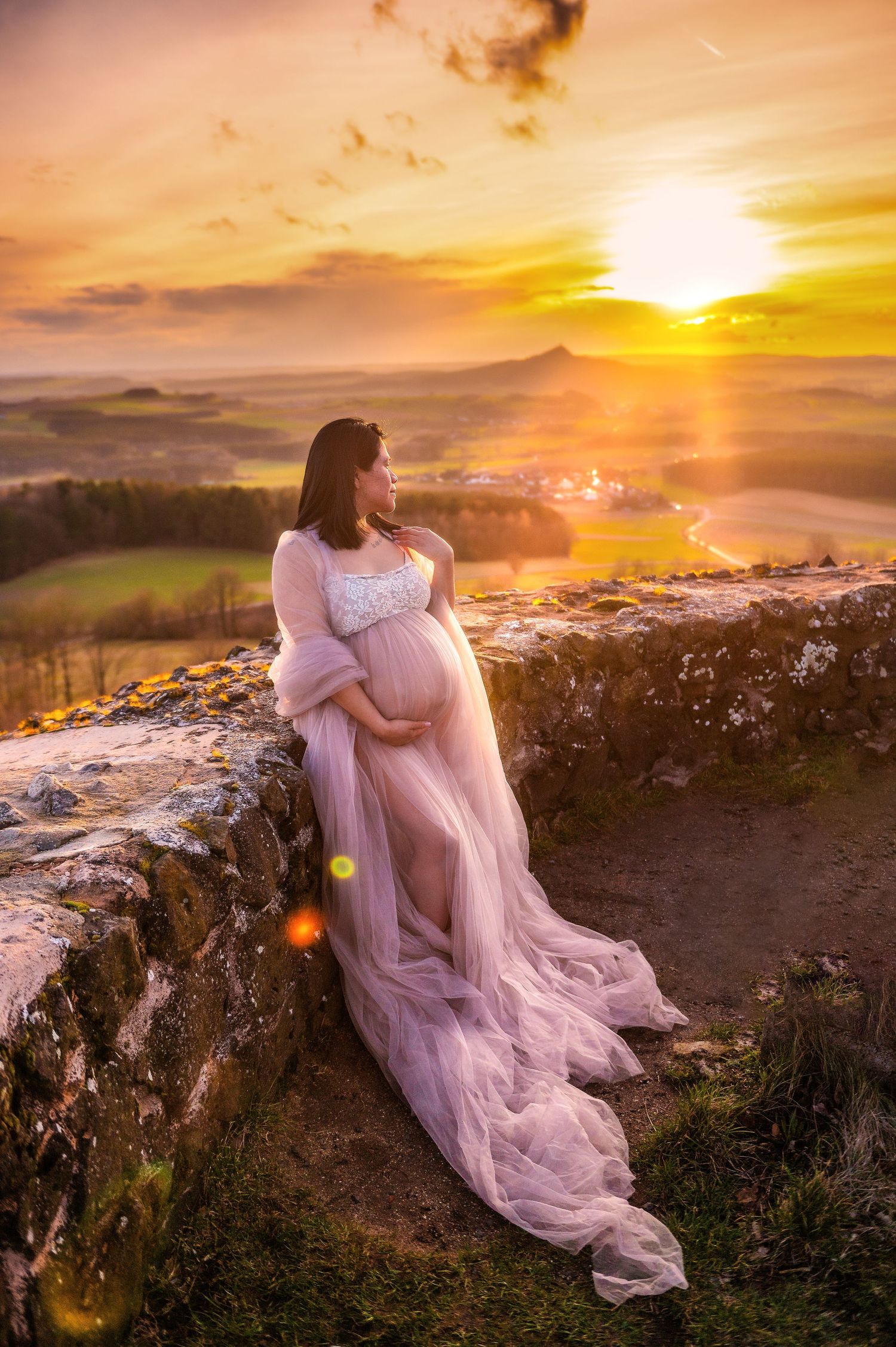 Ethereal maternity photo with flowing pink dress against golden sunset landscape.