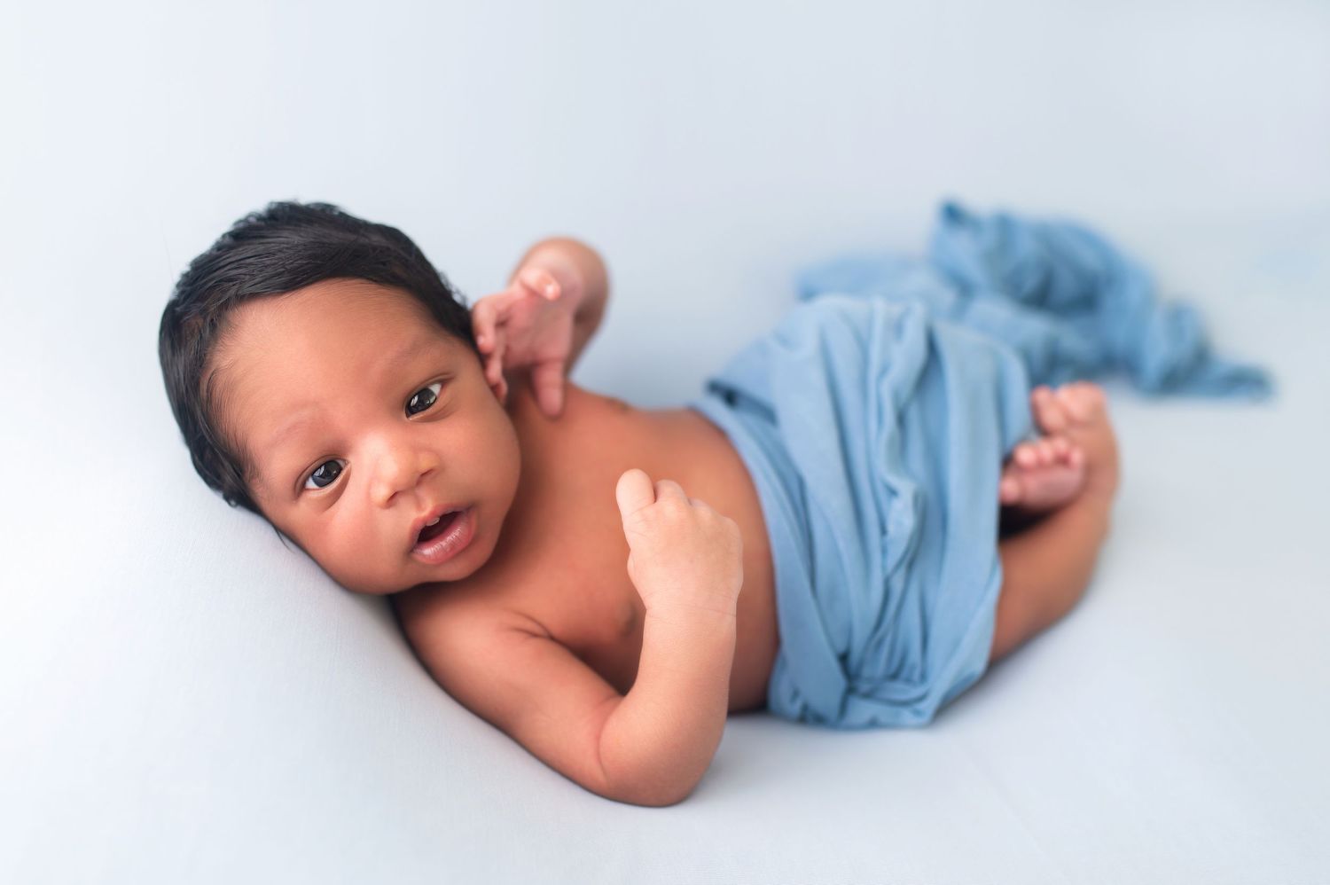 Baby in light blue wrap lying on white backdrop.
