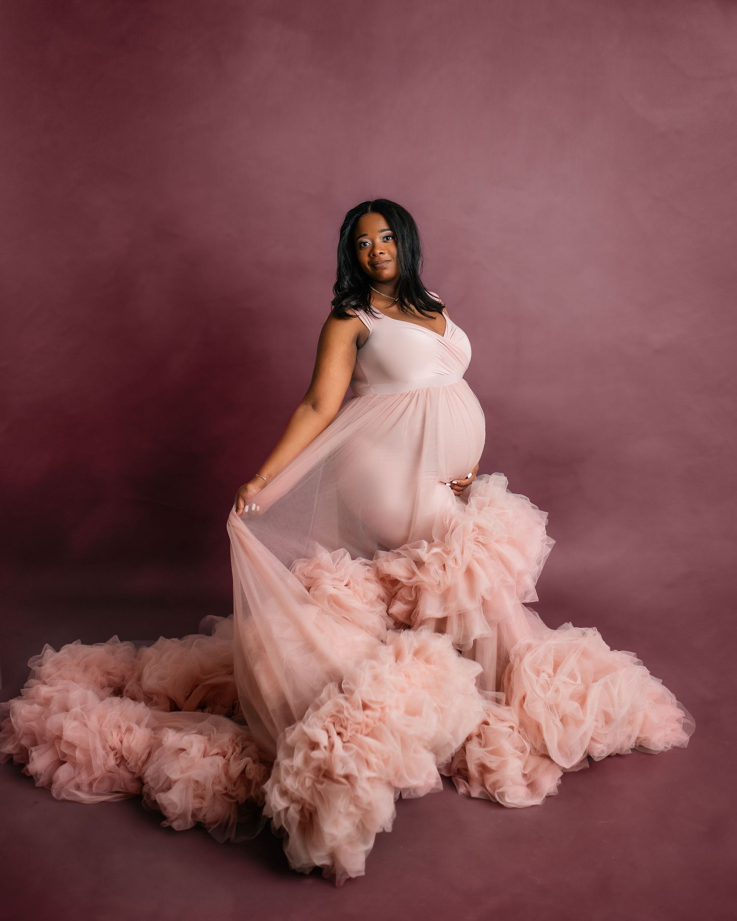 Pregnant woman in flowing pink tulle gown poses against burgundy backdrop in studio setting.