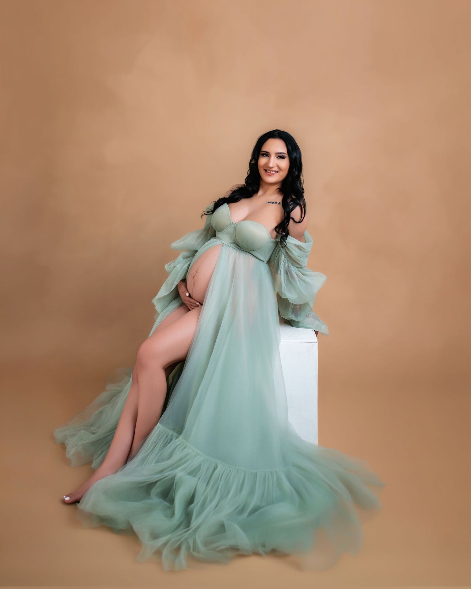 Pregnant woman in sage green tulle maternity gown posing elegantly on white cube against tan background.
