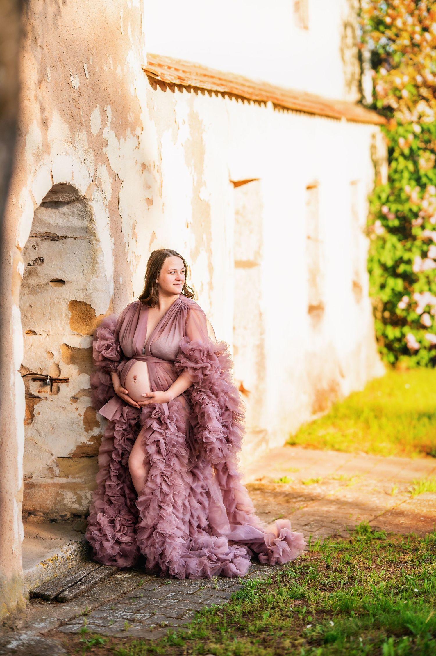 Maternity portrait in luxurious ruffled mauve gown against weathered stone wall and ivy.