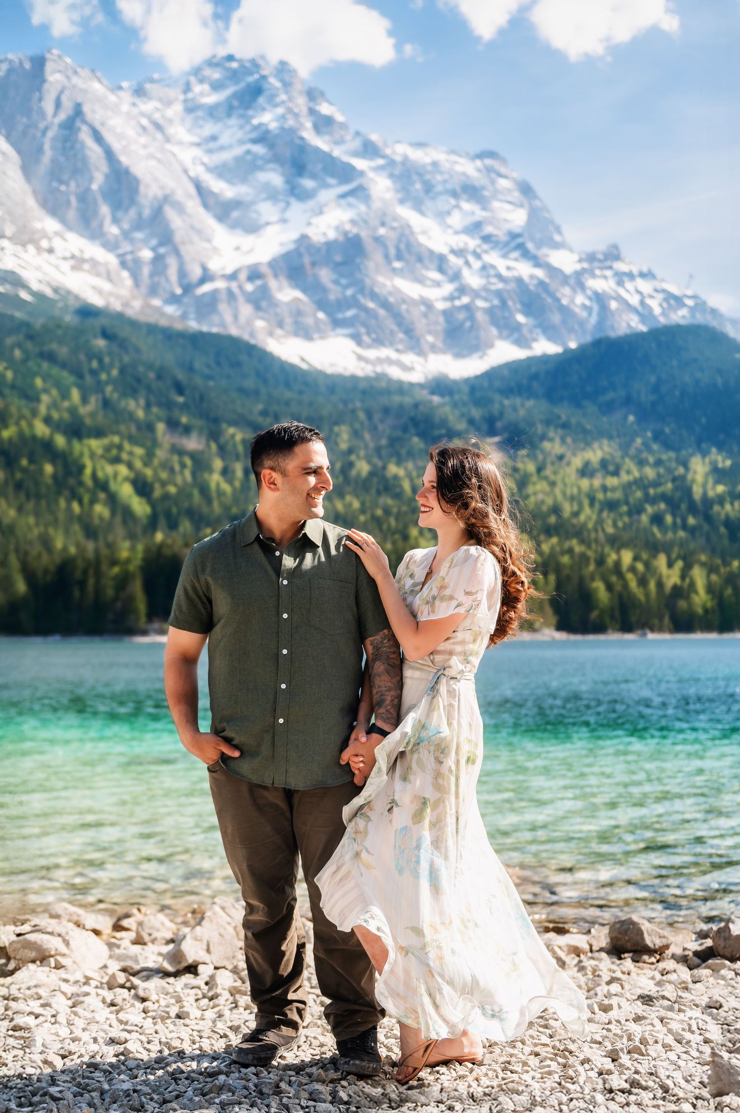 Couple holding hands by turquoise alpine lake with snow-capped mountains, woman in floral dress, man in green shirt.