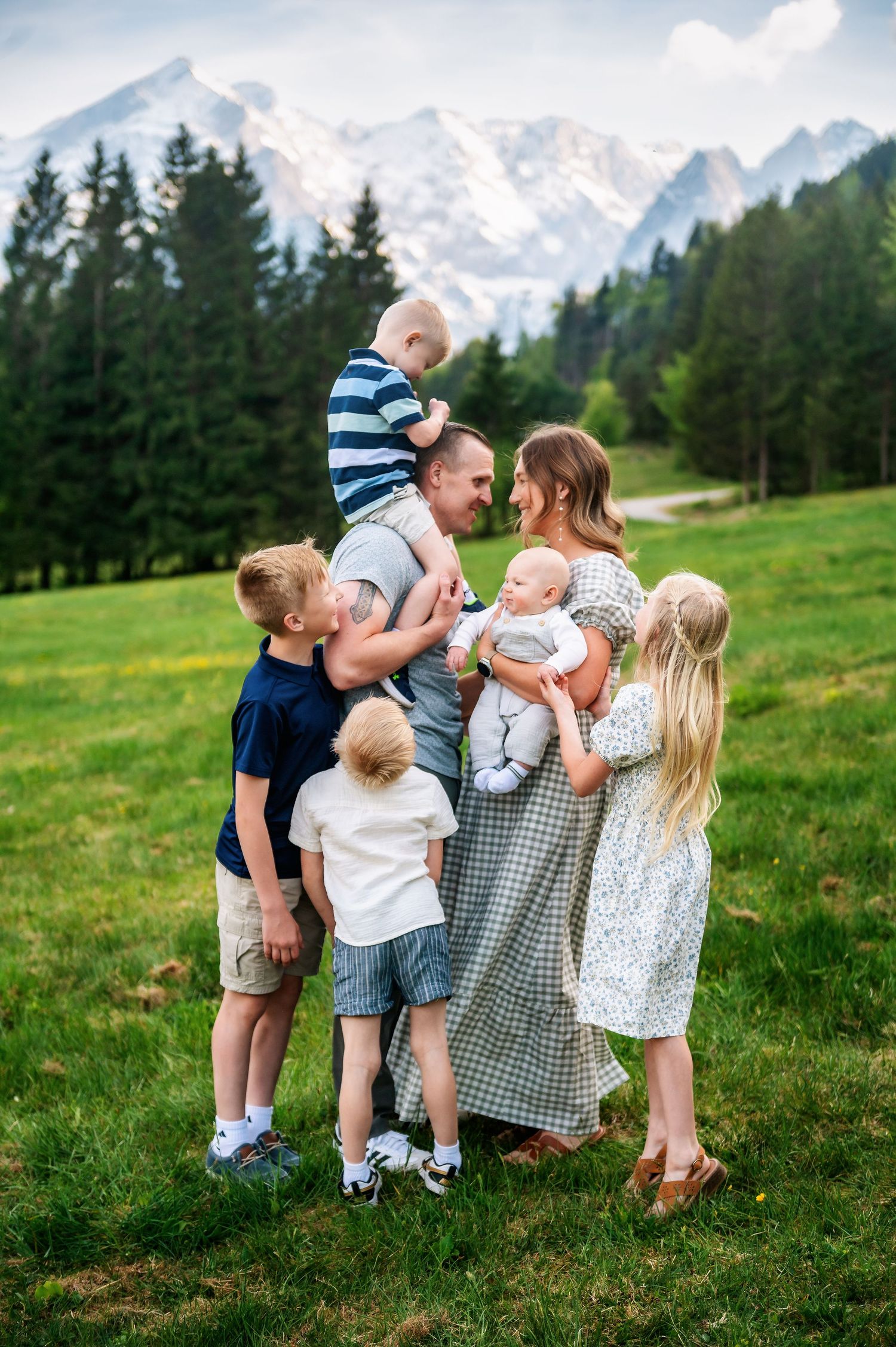 Family of seven posing together on green meadow with snow-capped mountains and pine trees in background.