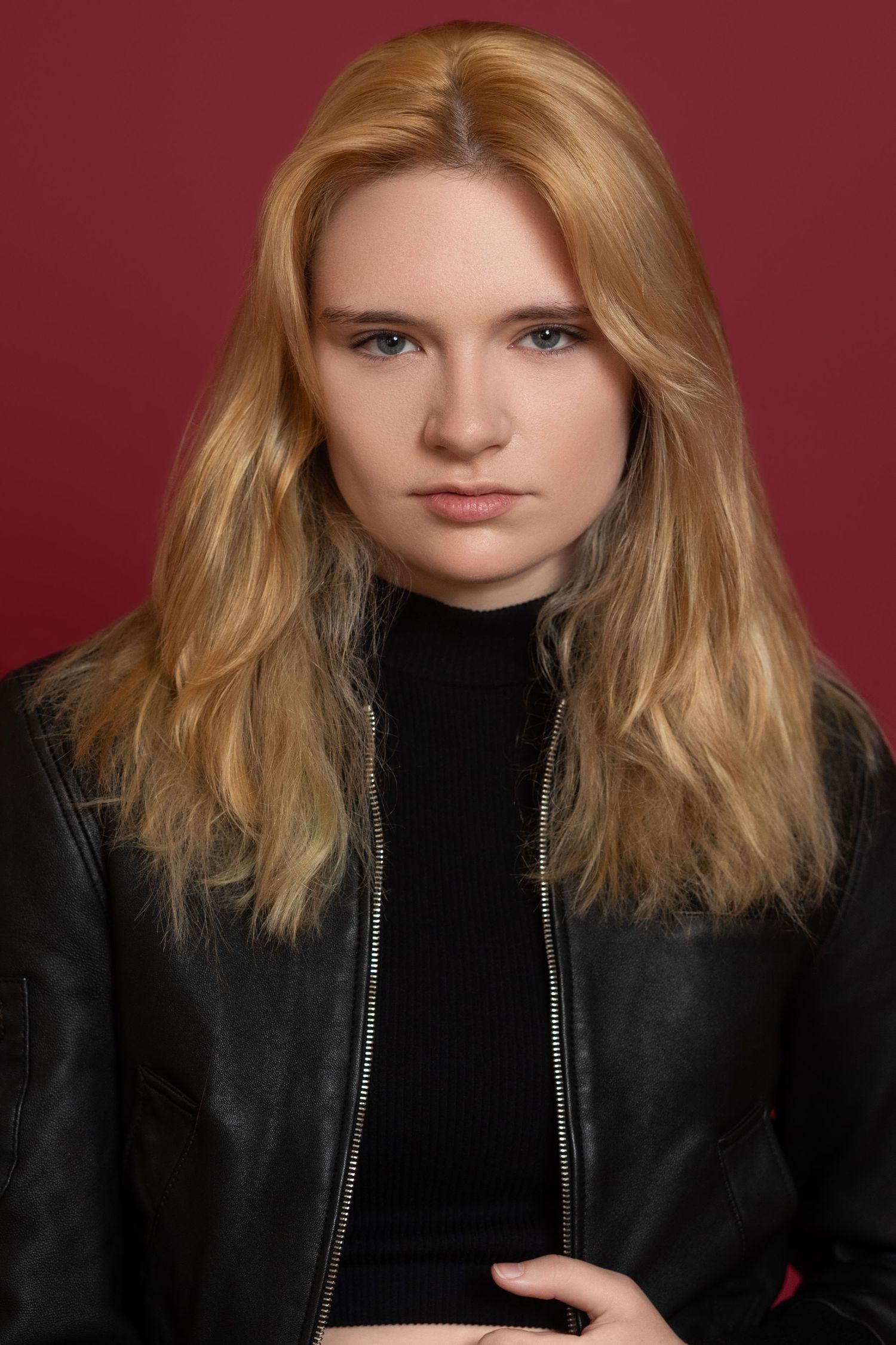 Studio portrait against burgundy background showing person in black turtleneck and leather jacket.