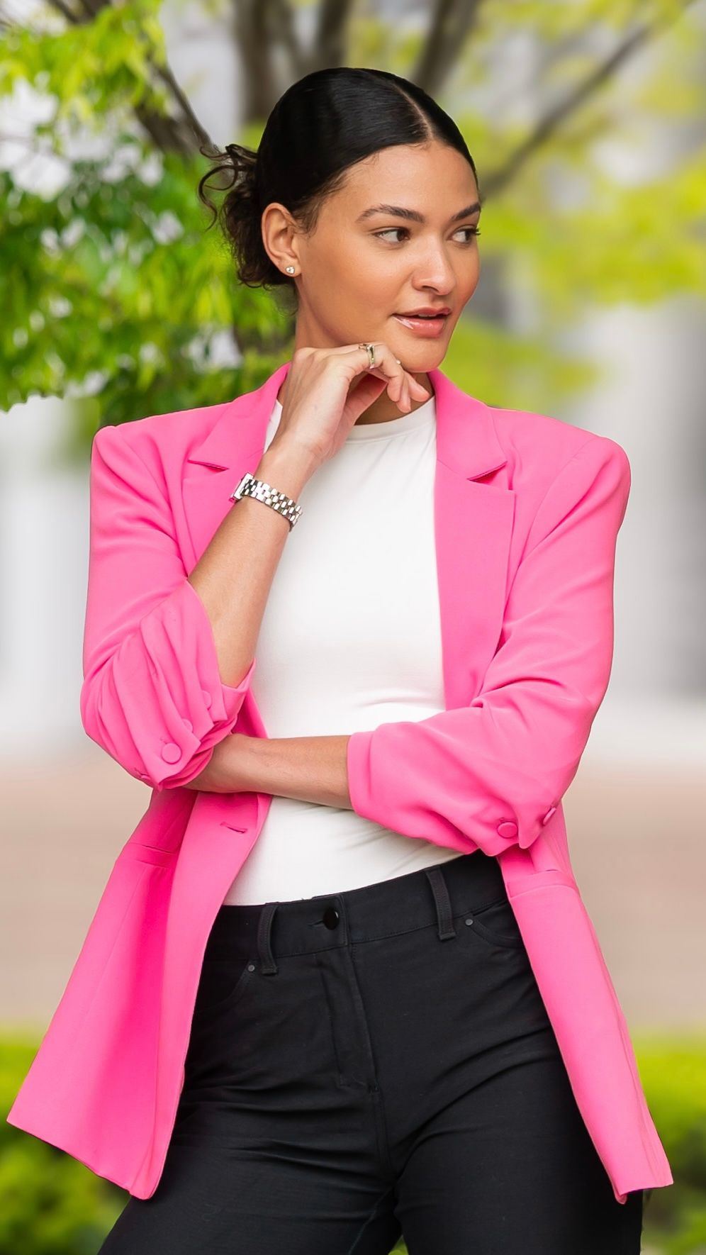 Fashion model wearing bright pink blazer with white top and black pants in outdoor setting with soft natural lighting.