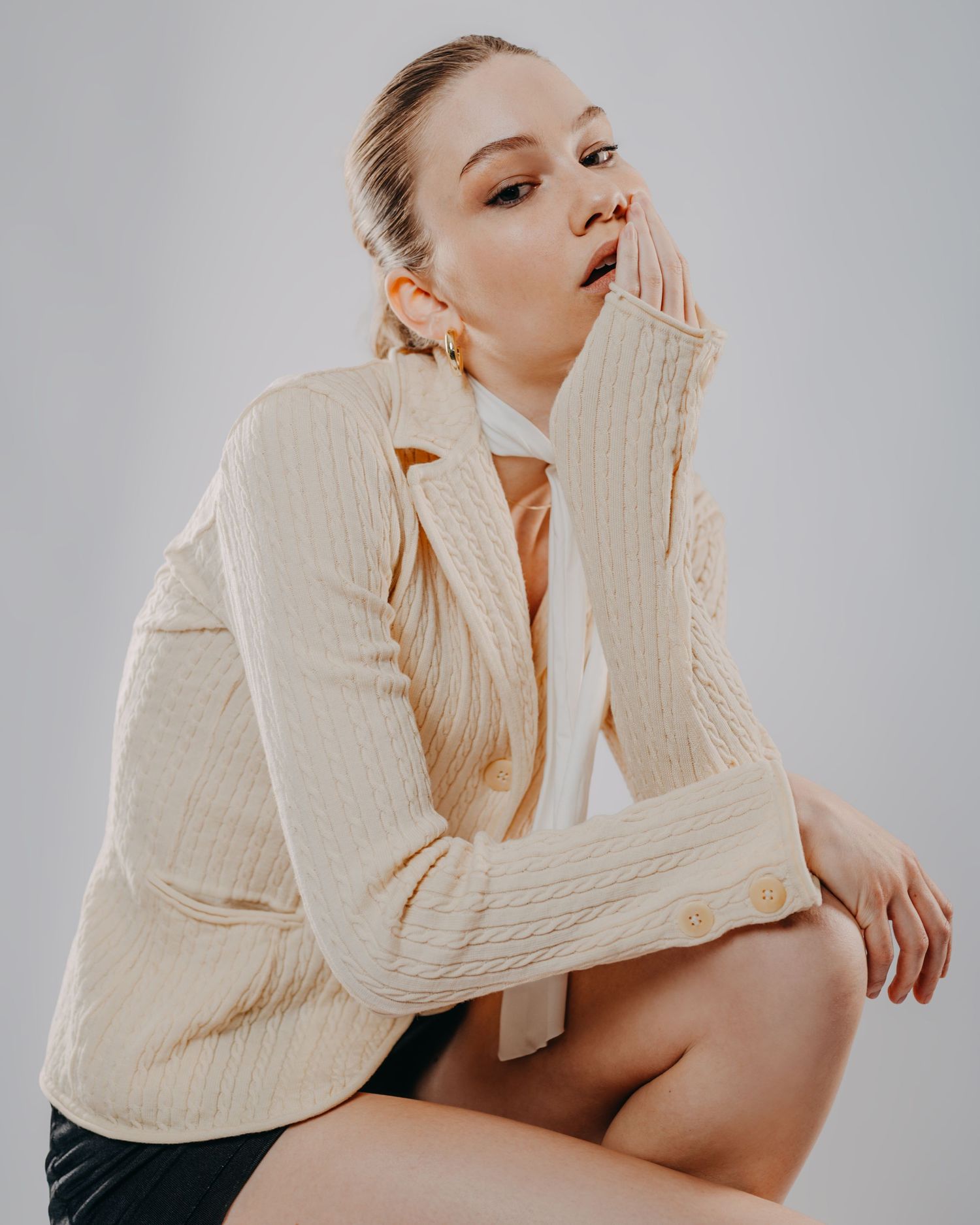 Fashion model in cream cable knit sweater poses thoughtfully with hand near face.