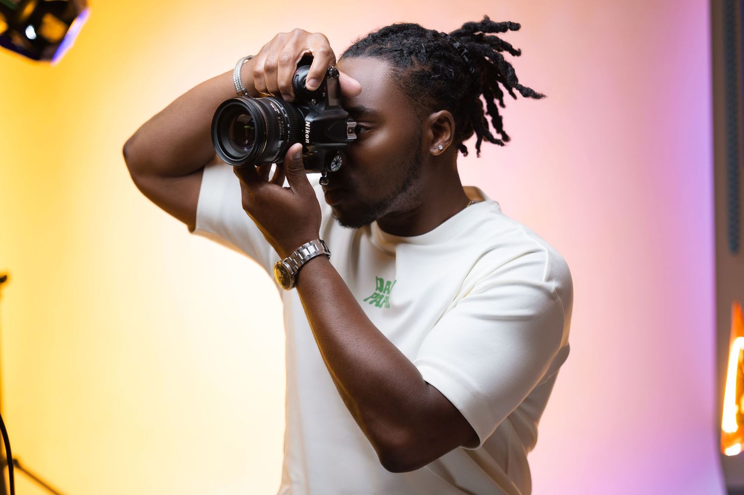 A photographer wearing a white t-shirt holds up a camera to take a photo against a yellow and purple gradient background.