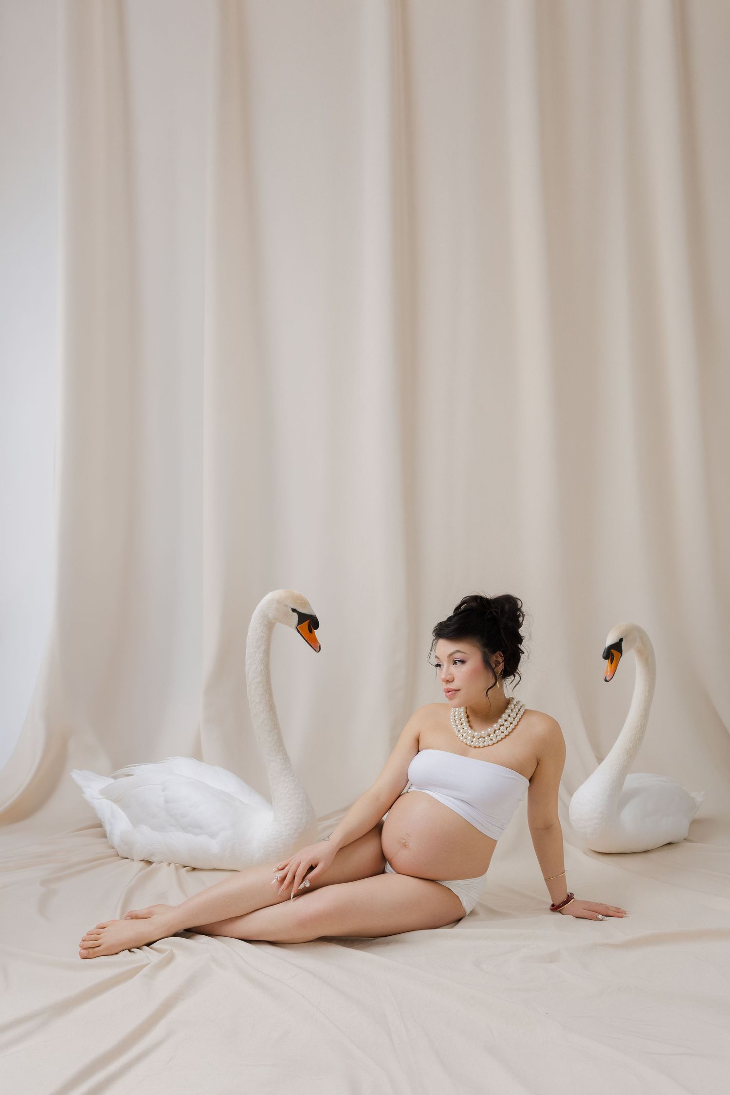 Pregnant woman in white bandeau sits gracefully between two white swans in a serene, neutral-toned studio setting.