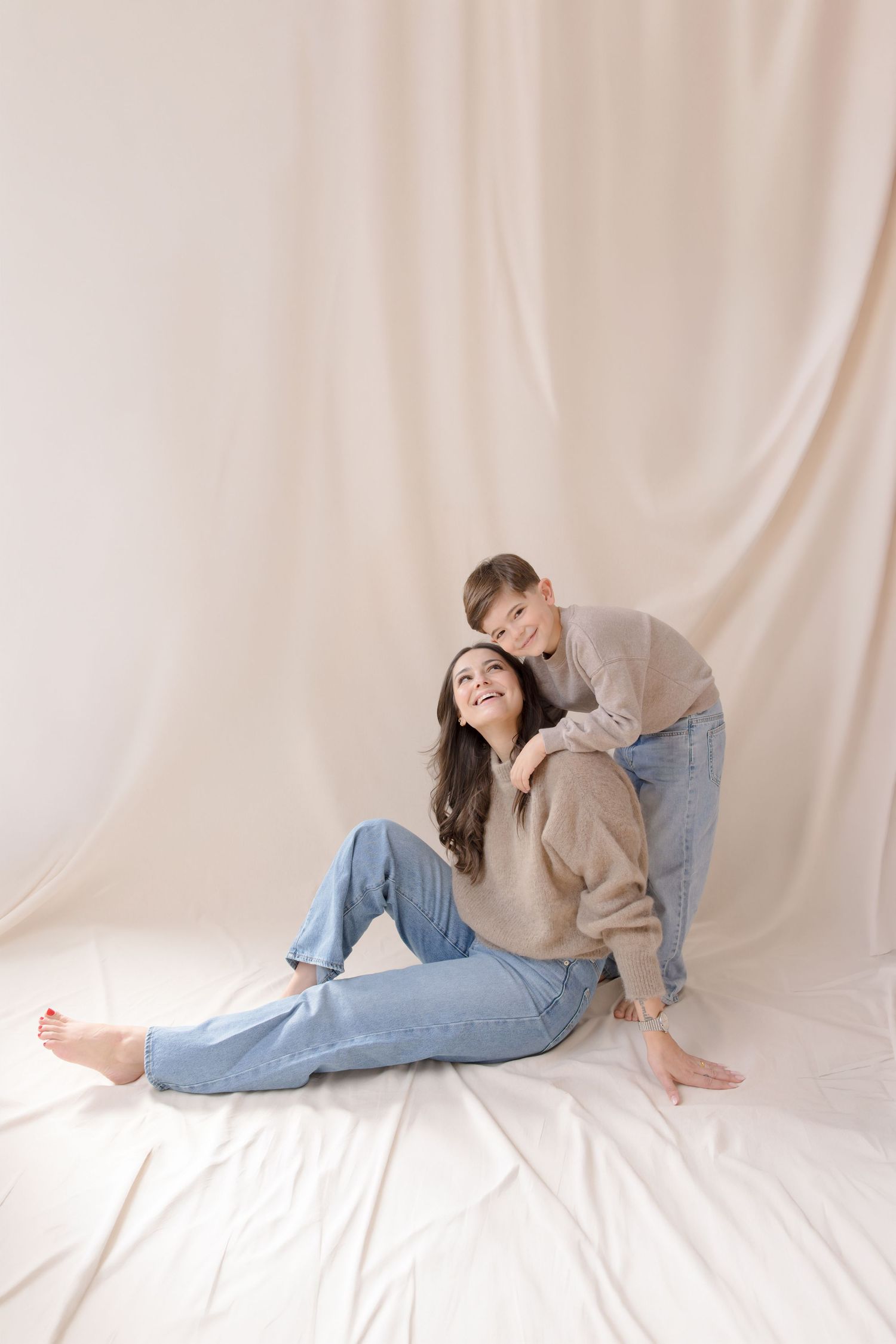 A woman sits on the floor smiling as a child leans over her shoulder, both wearing beige sweaters and blue jeans.