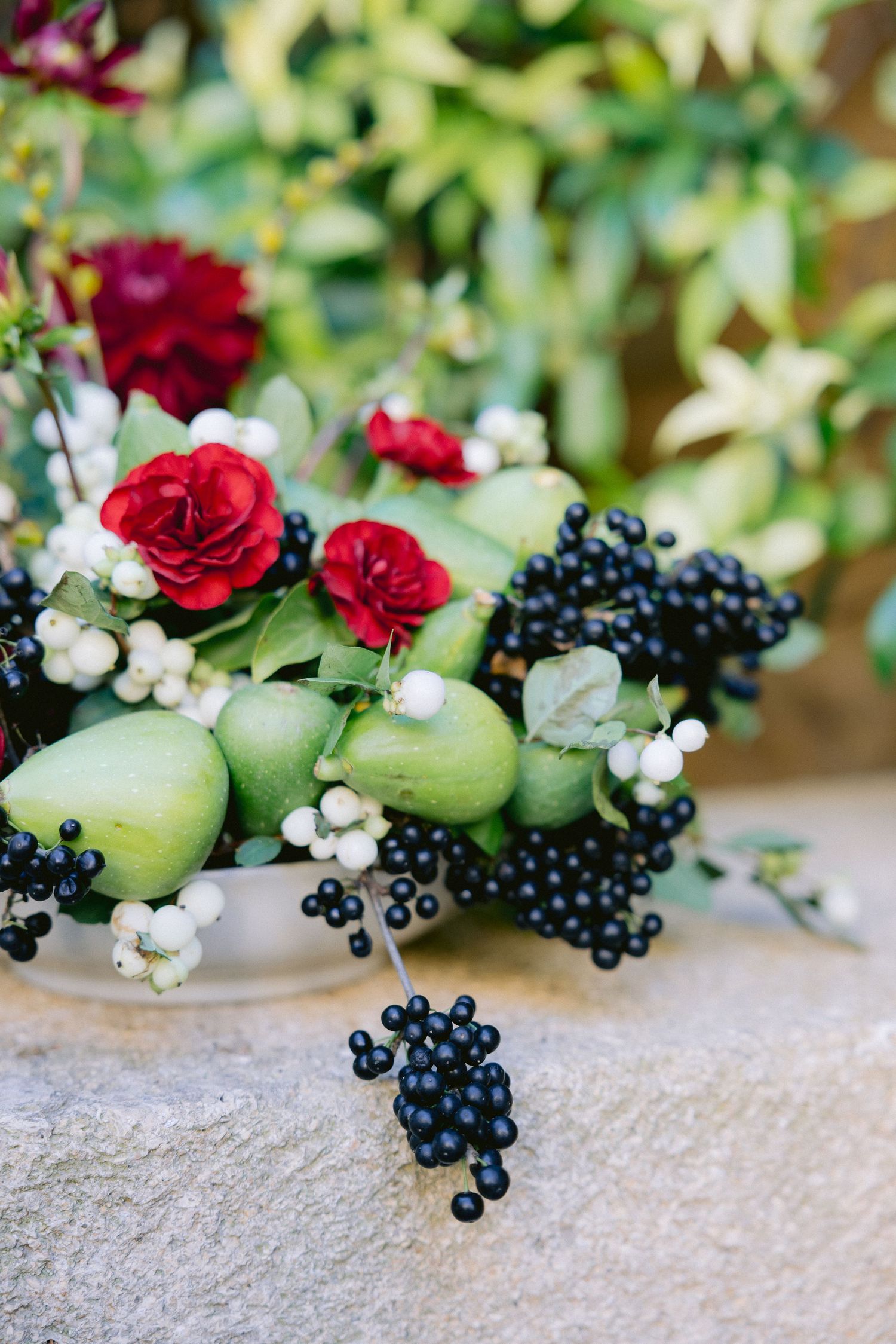 Decorative floral arrangement with red roses, green apples and black berries on stone surface.