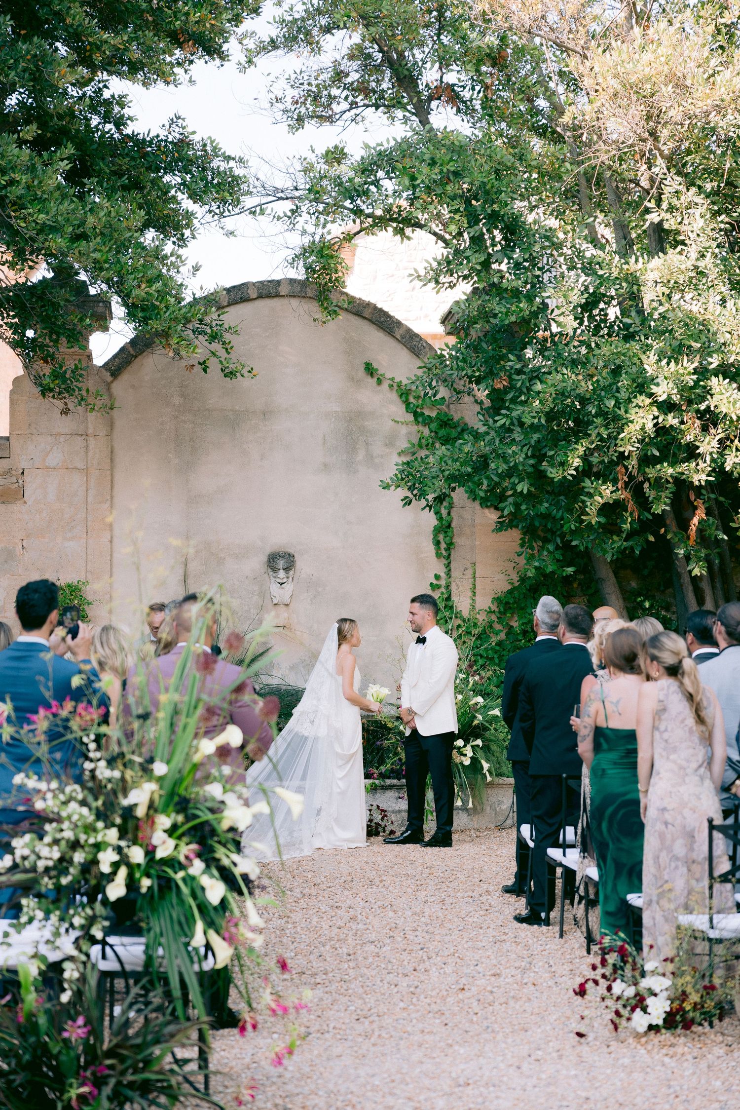 Wedding ceremony in progress in courtyard with stone walls and natural greenery decorations.