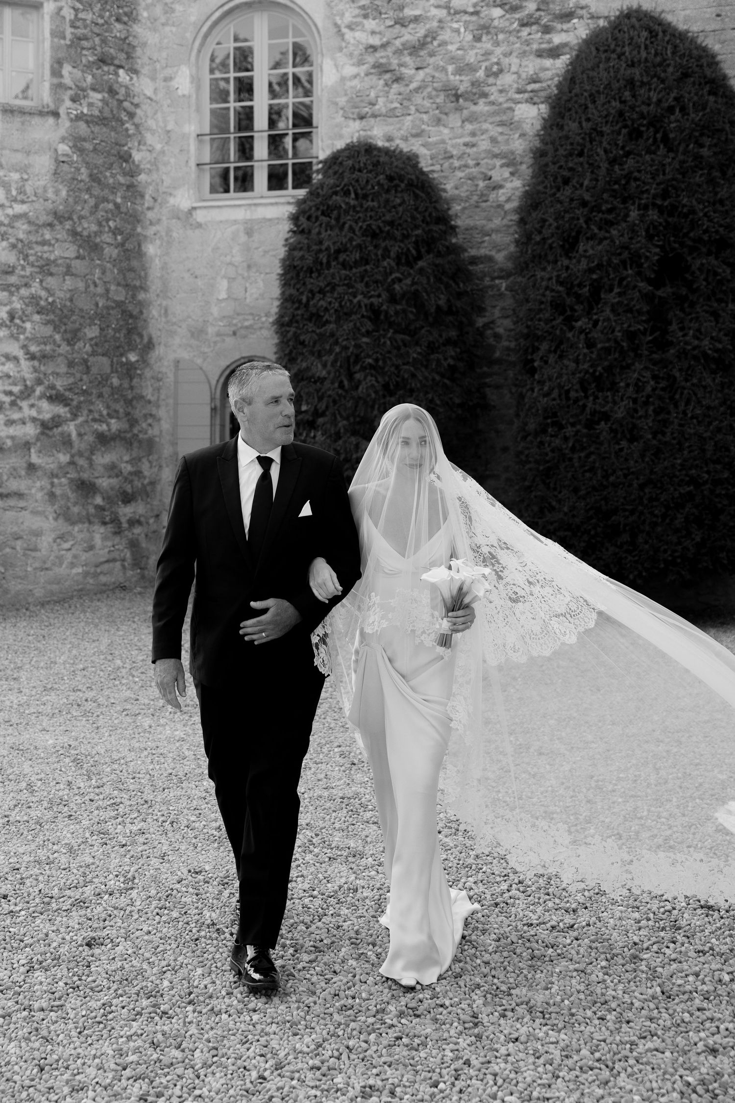 Black and white wedding photos of a couple walking together on gravel path near stone building.