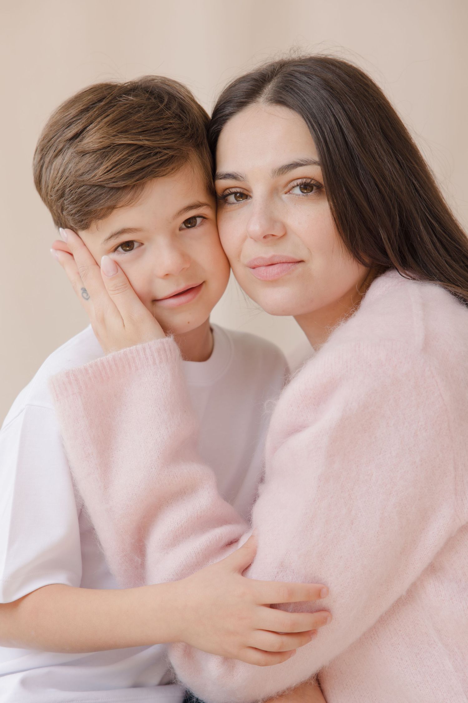A young boy and dark-haired woman in a pink sweater share a warm embrace, posing together for a portrait photo.
