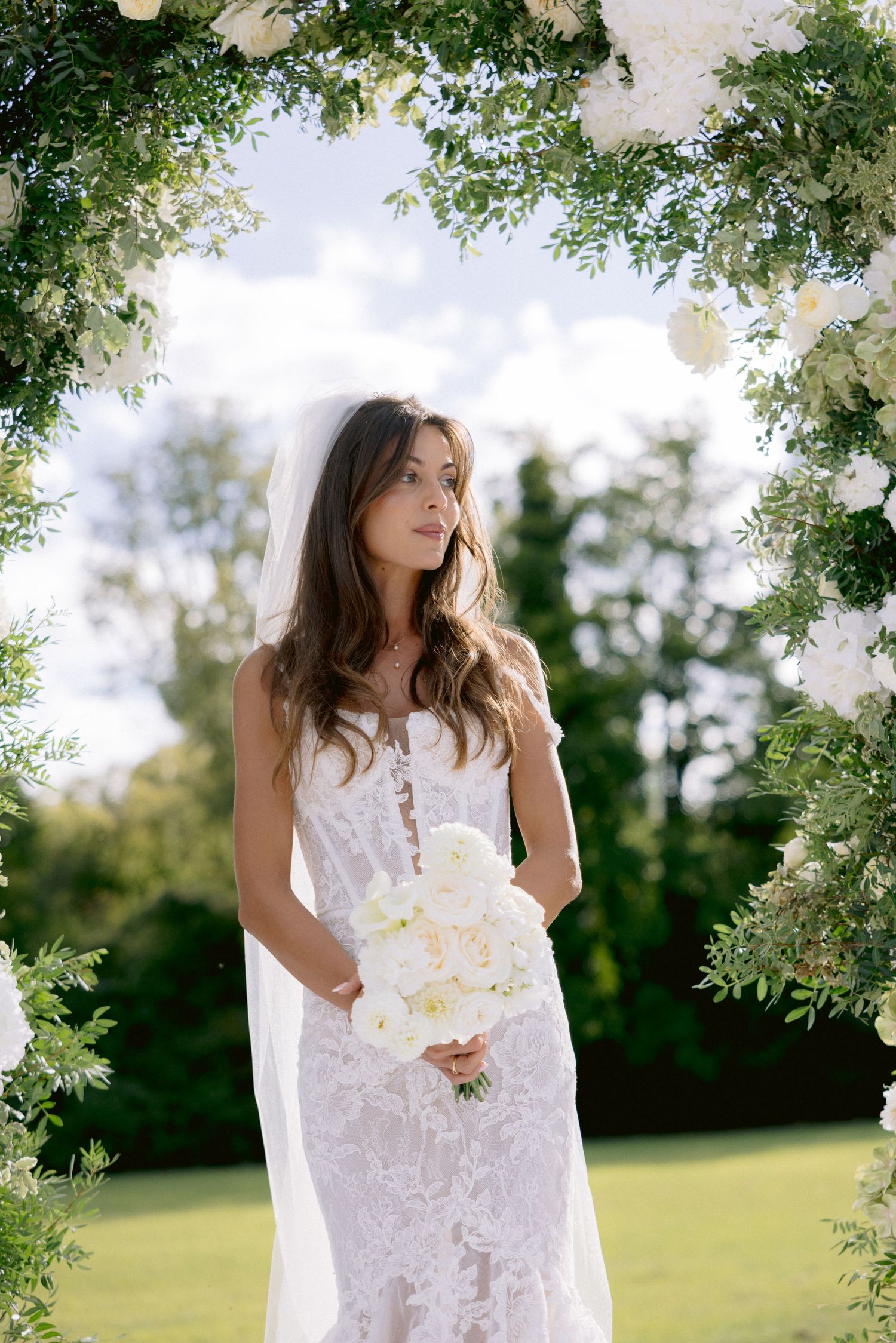 Elegant outdoor wedding arch decorated with white flowers and greenery against a sunny landscape.