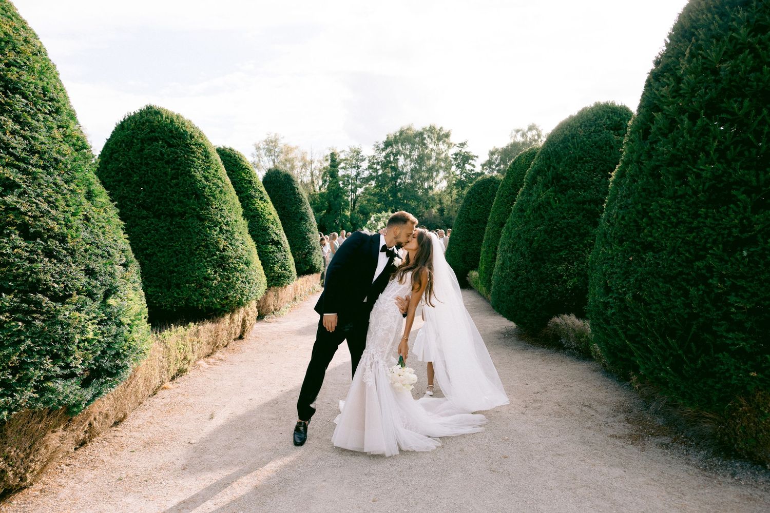 Wedding couple walking through a manicured garden path surrounded by tall hedges.