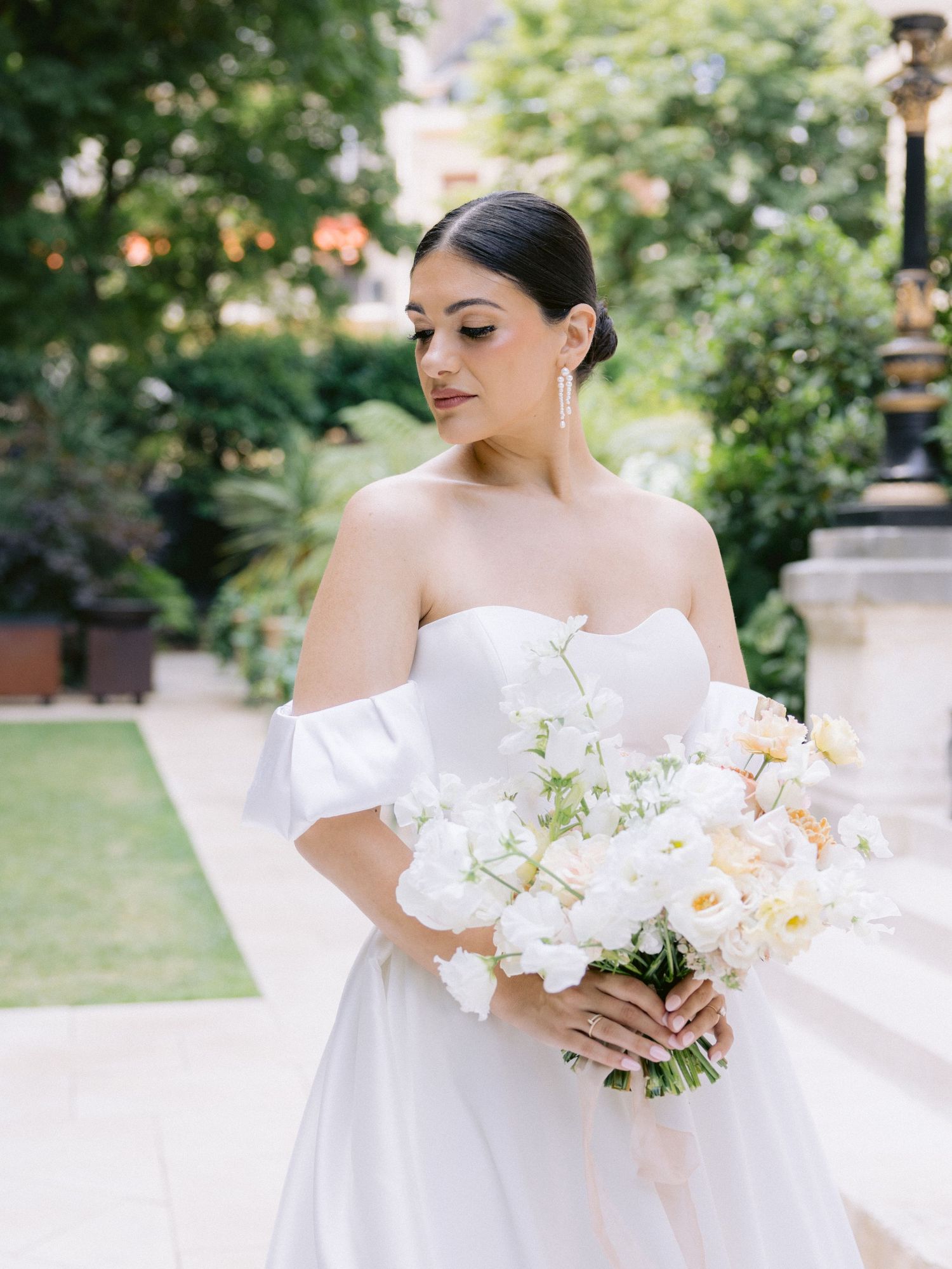 Elegant bride in off-shoulder white gown holding delicate bouquet of white and peach flowers.
