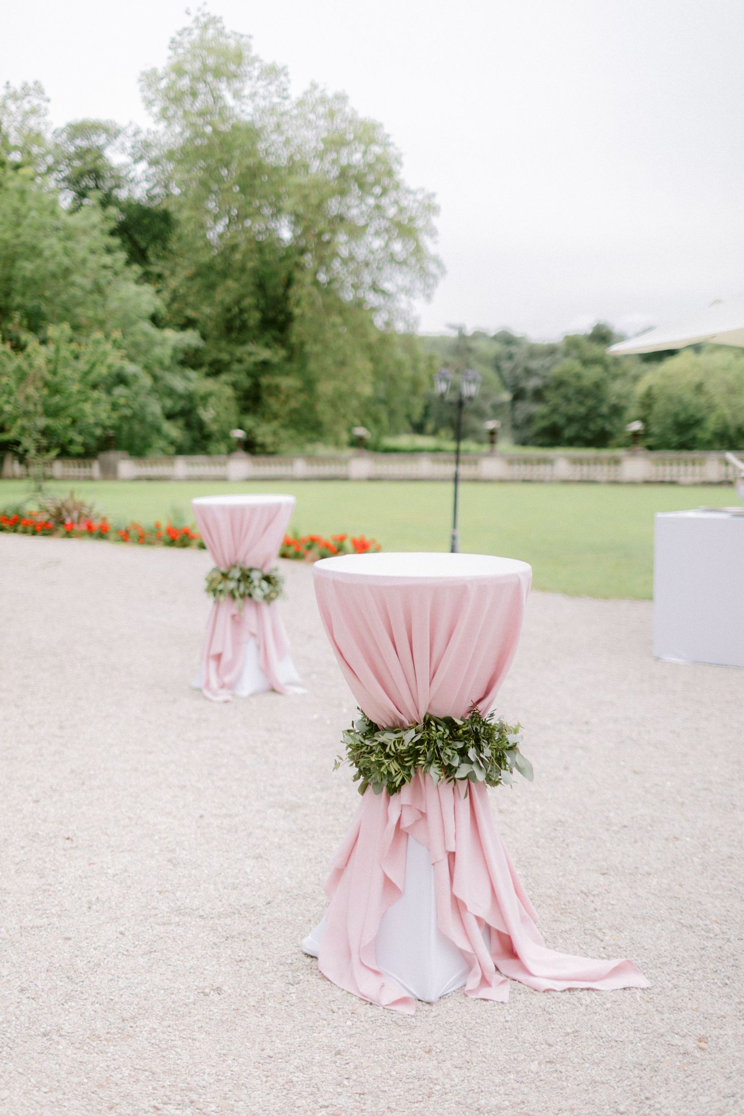 Pink draped cocktail tables decorated with greenery garlands set up outdoors for an event.