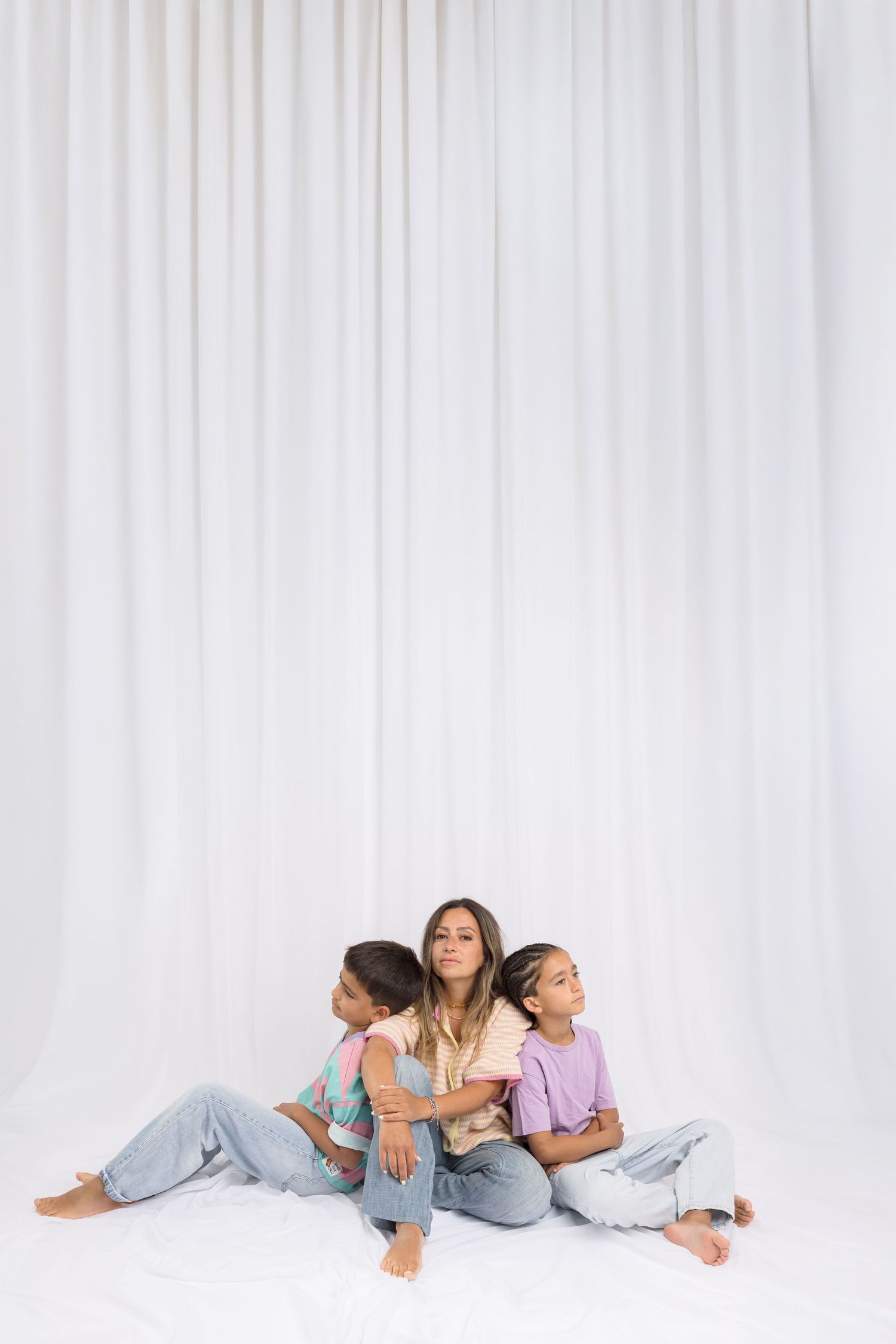 A woman sits on the floor with two children leaning against her, posed in front of a white curtain backdrop.