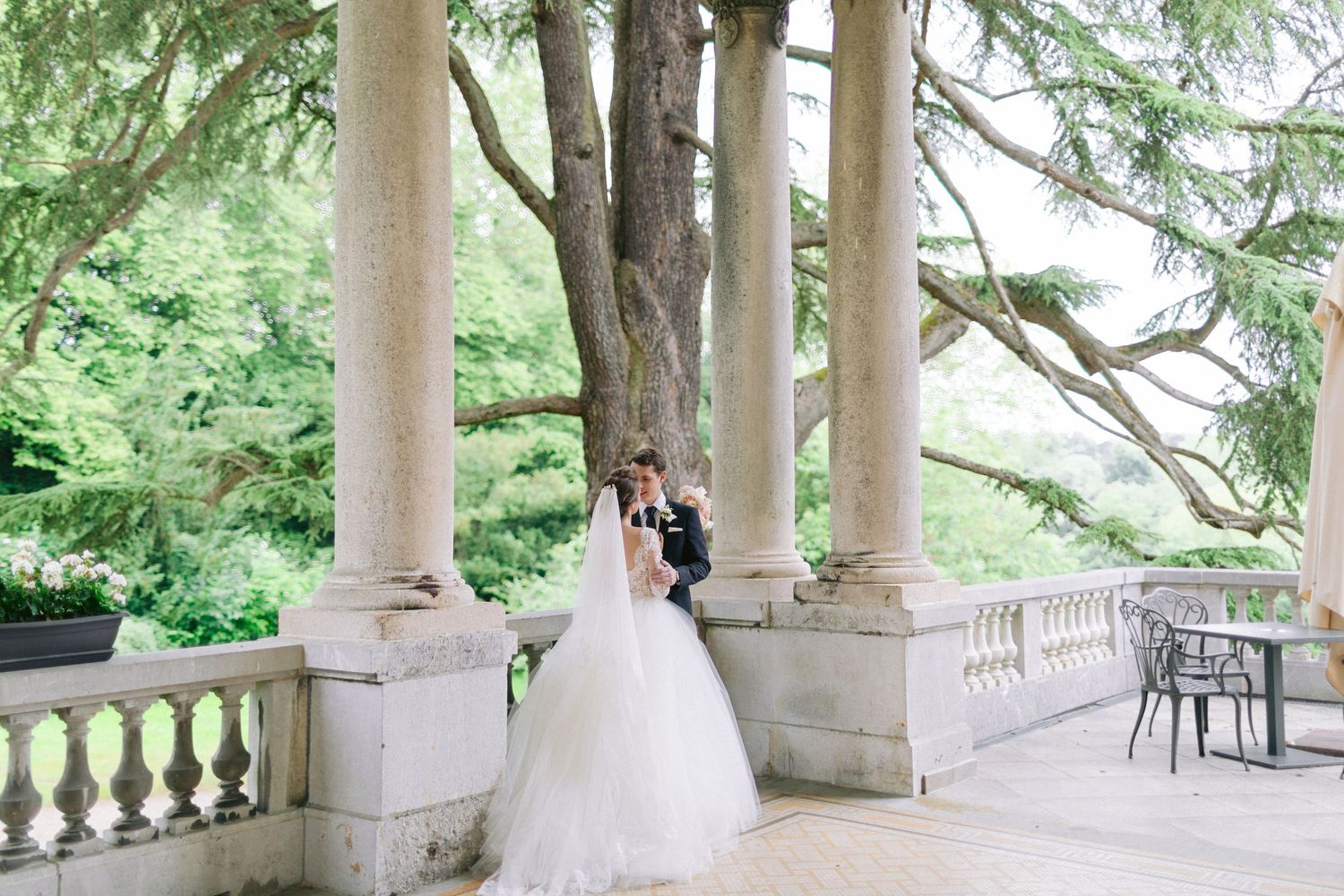 A wedding couple standing between classic stone columns surrounded by lush greenery.