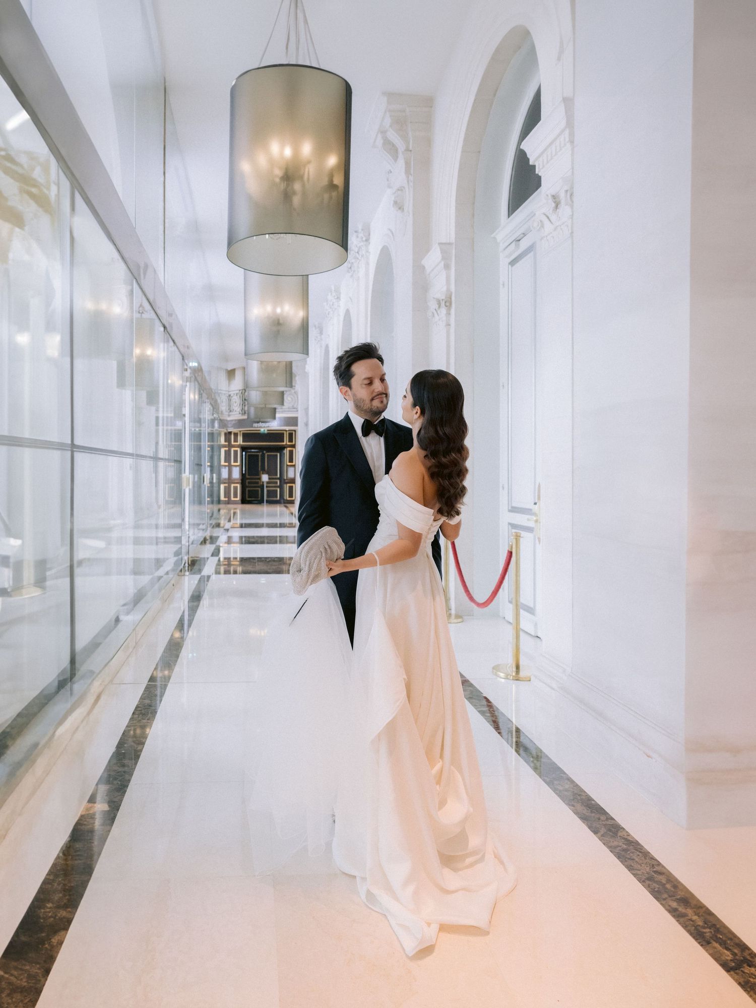 Elegant couple walks through a bright white hallway in formal wedding attire.