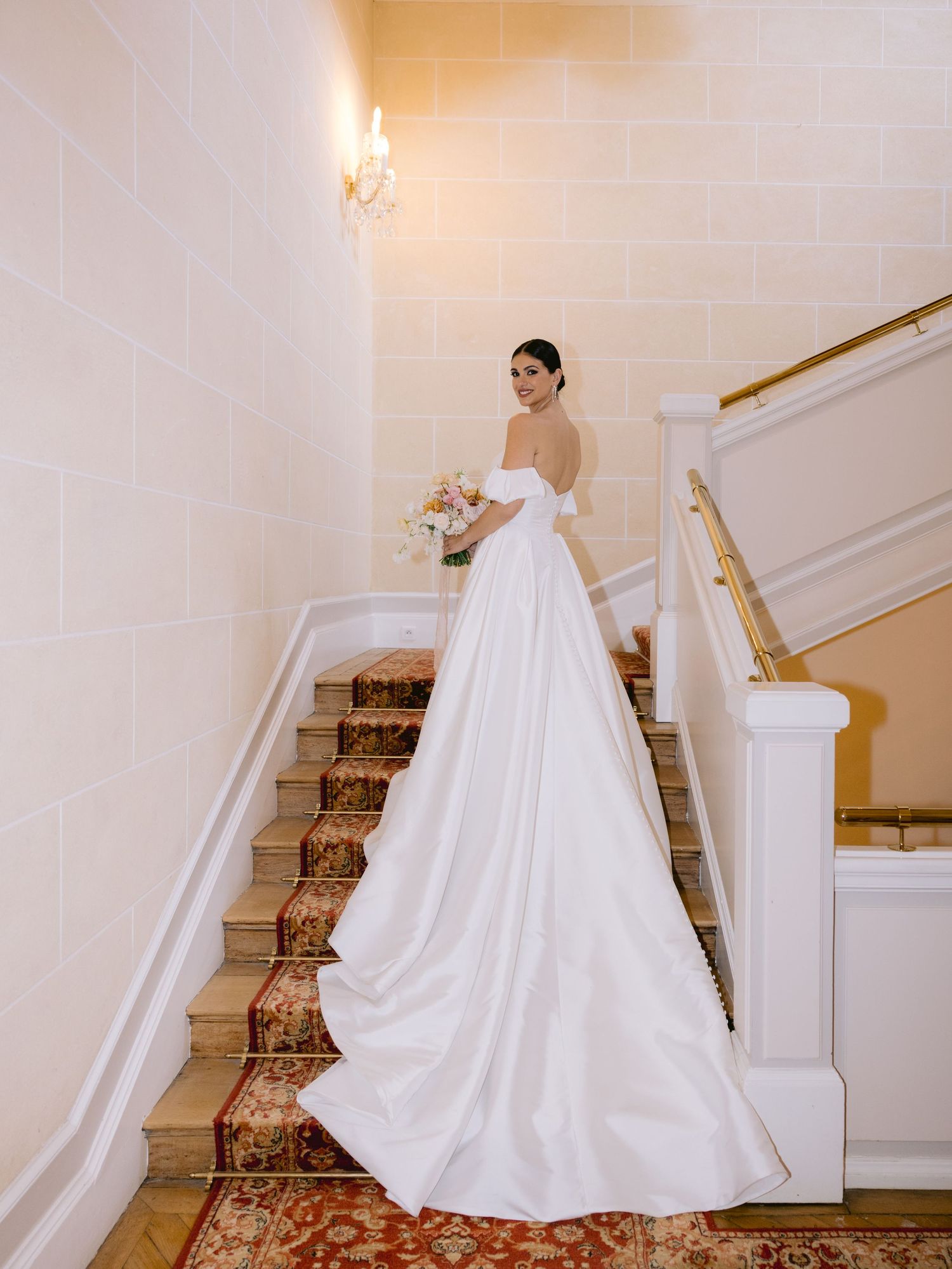 A flowing white wedding gown with a dramatic train cascades down an elegant staircase with floral carpeting.
