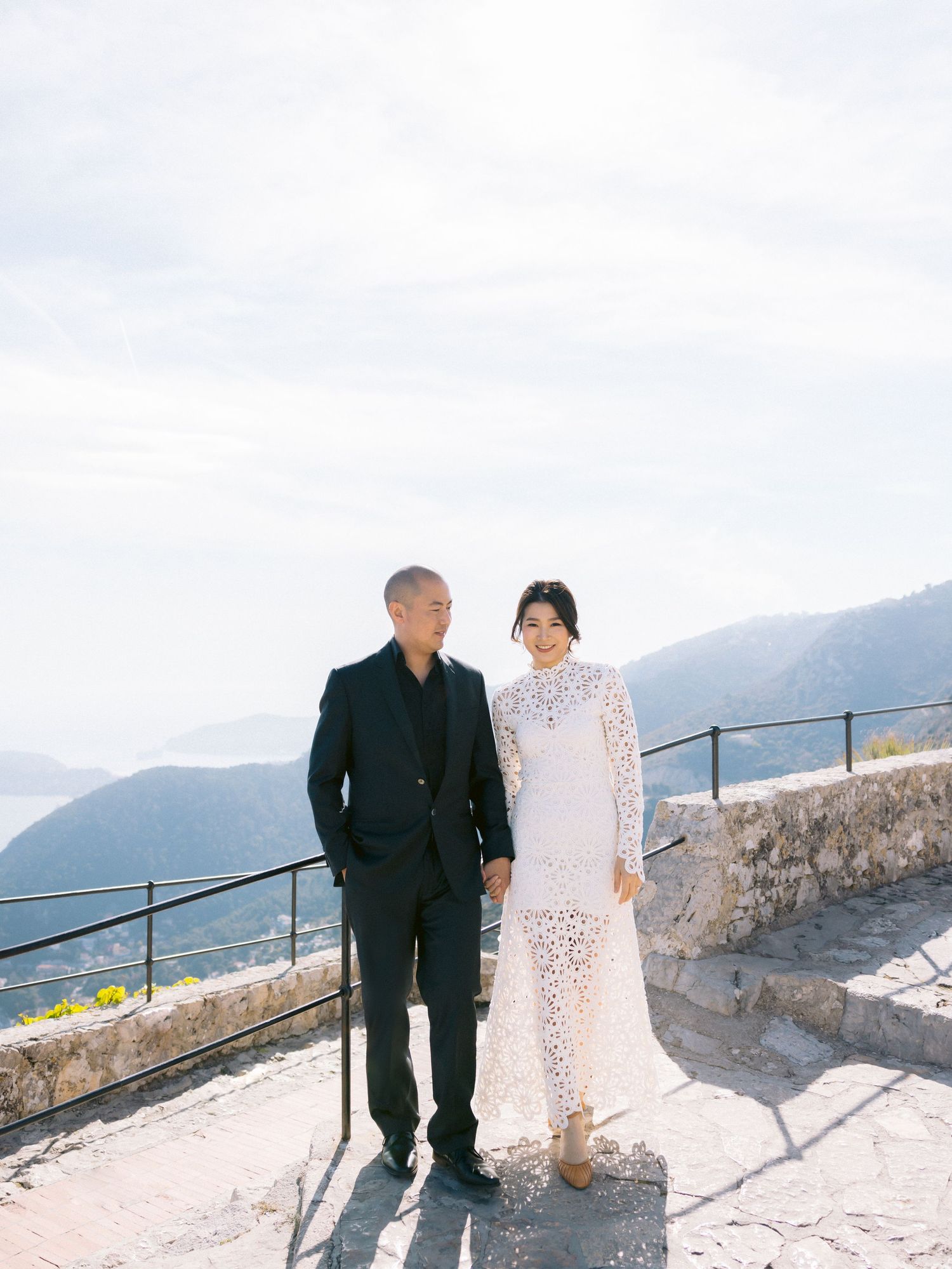 A couple in wedding attire stands together on a stone terrace overlooking a scenic mountain landscape on a sunny day.