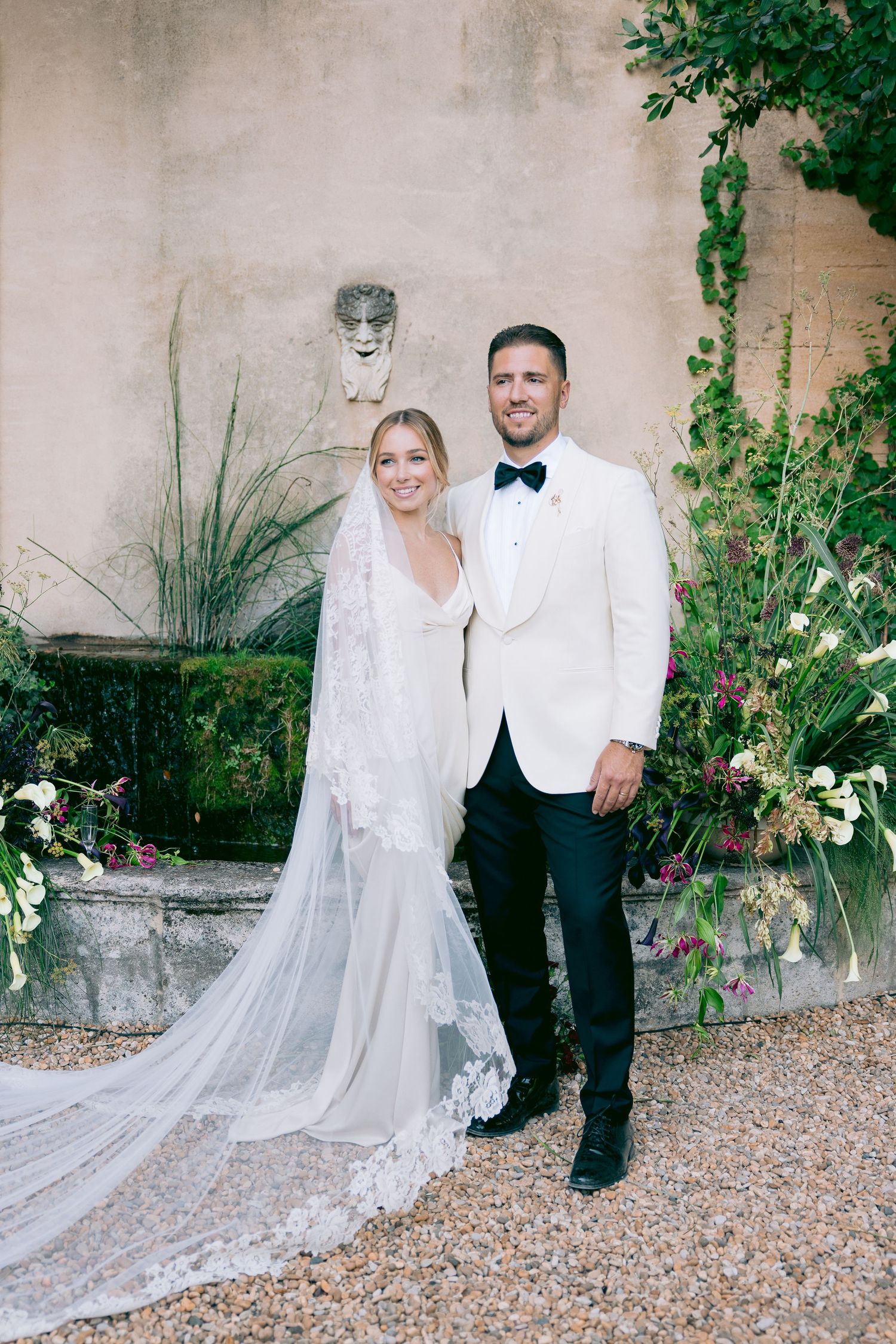 Elegant wedding couple posing against stone wall with flowing veil and formal attire.